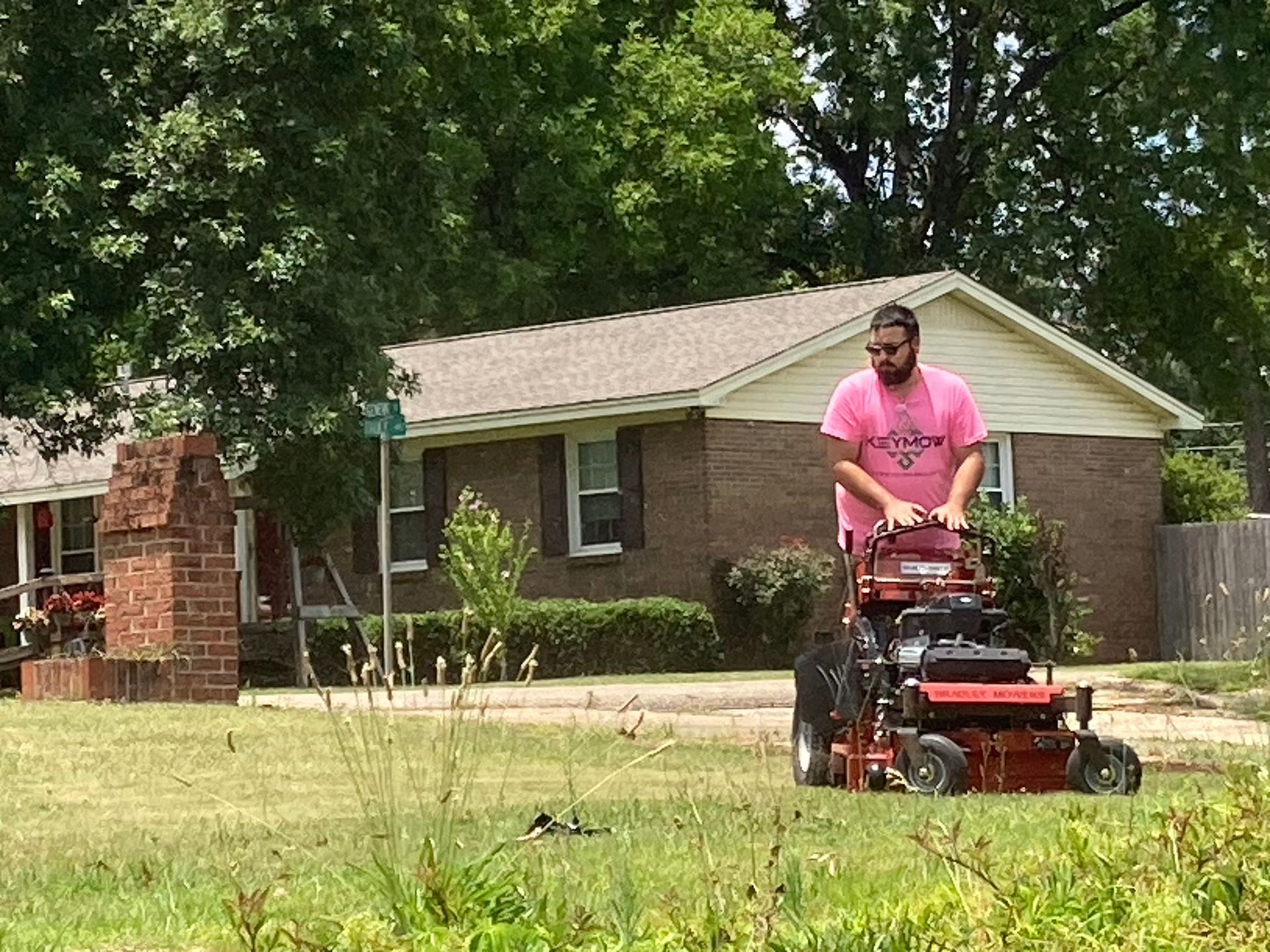 Person mowing a lawn with a red riding mower in front of a brick house