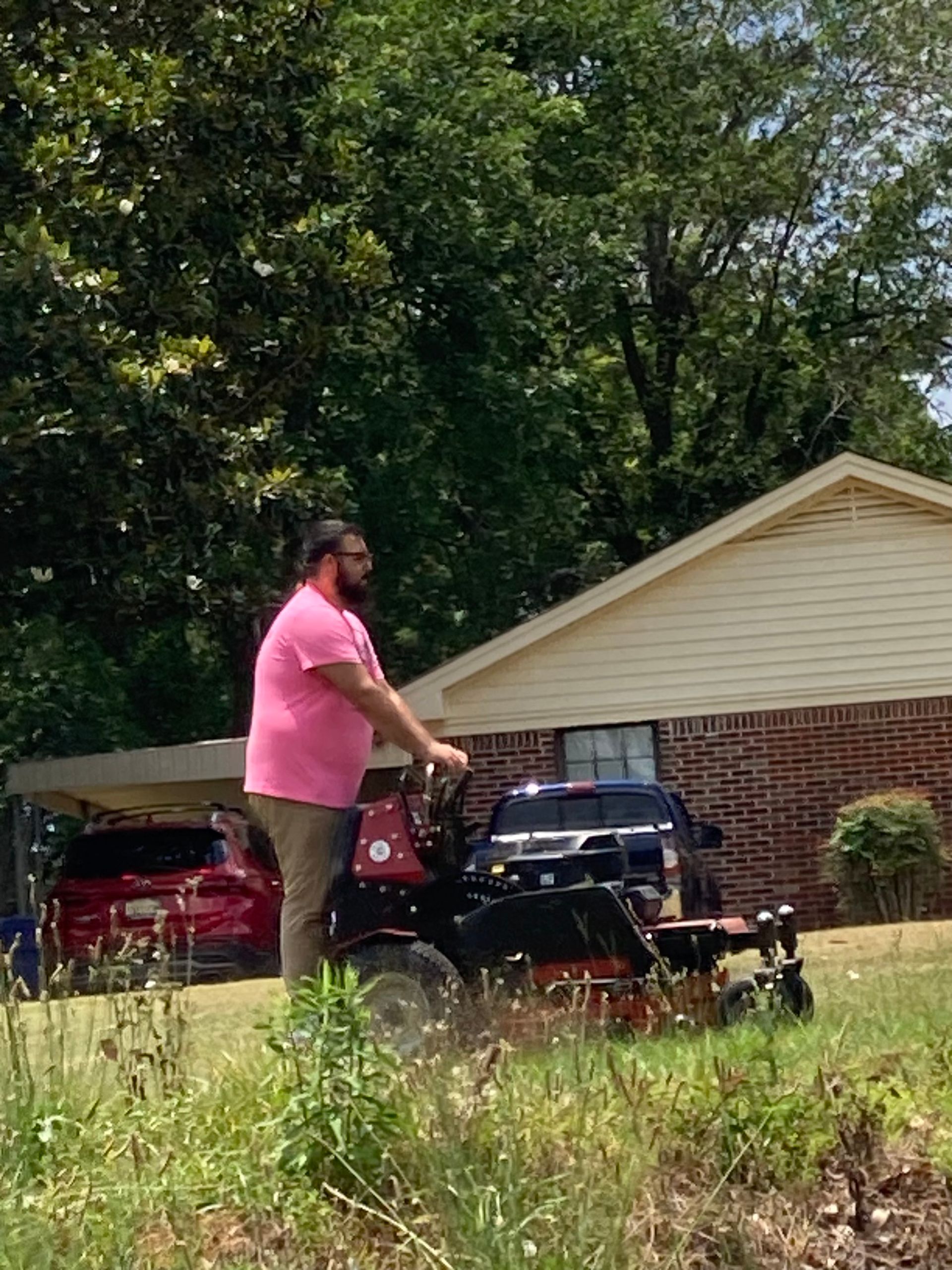 Person in pink shirt pushing a black lawn mower in a front yard with a brick house and trees.
