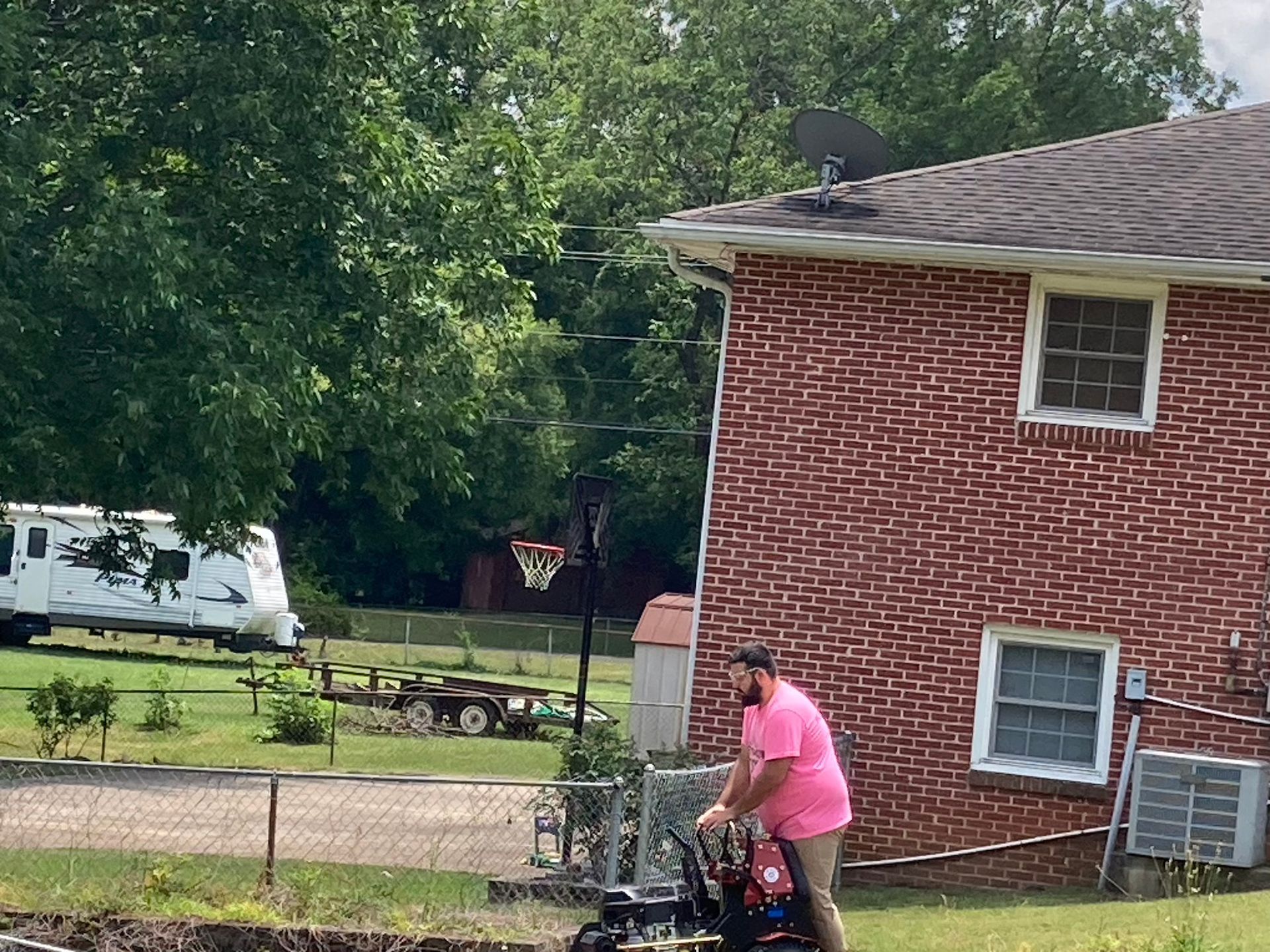 Person in a pink shirt mowing grass beside a red brick house, with trees and a parked RV in the background