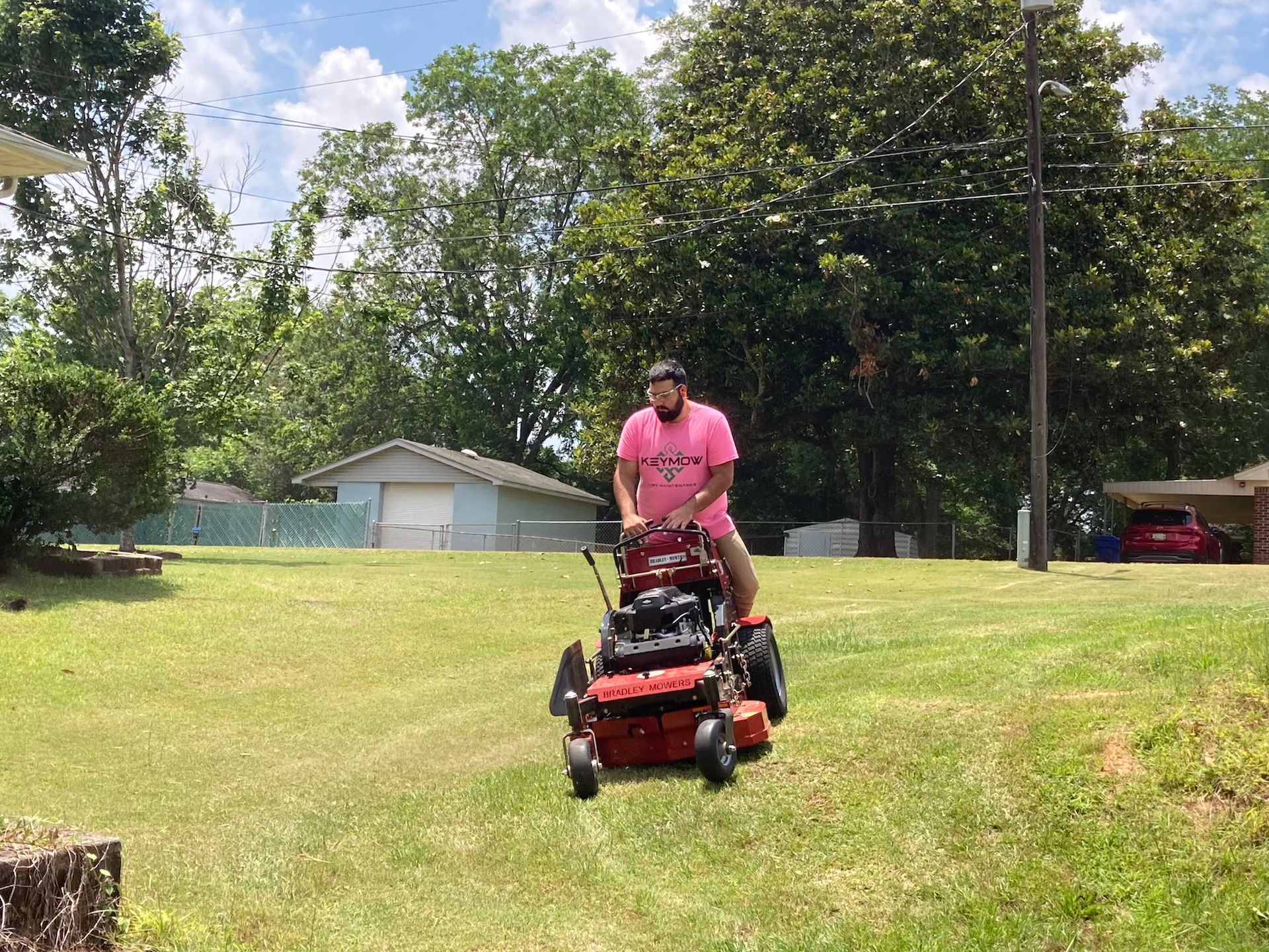 Person mowing a grassy lawn with a red riding mower near a house and trees