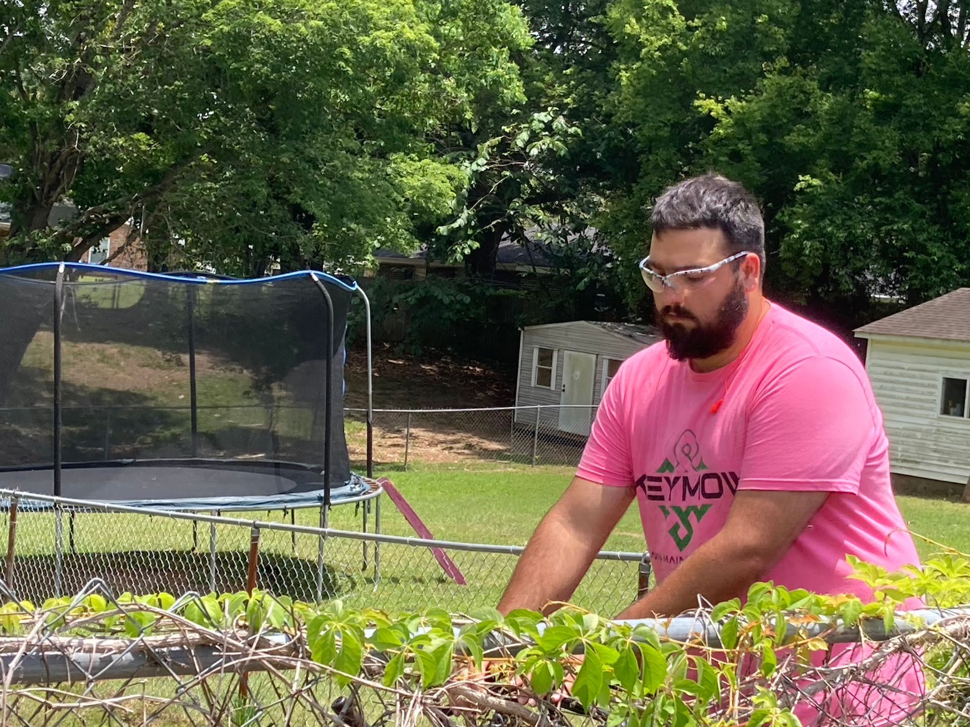 Man in a pink shirt tending a garden beside a trampoline in a backyard