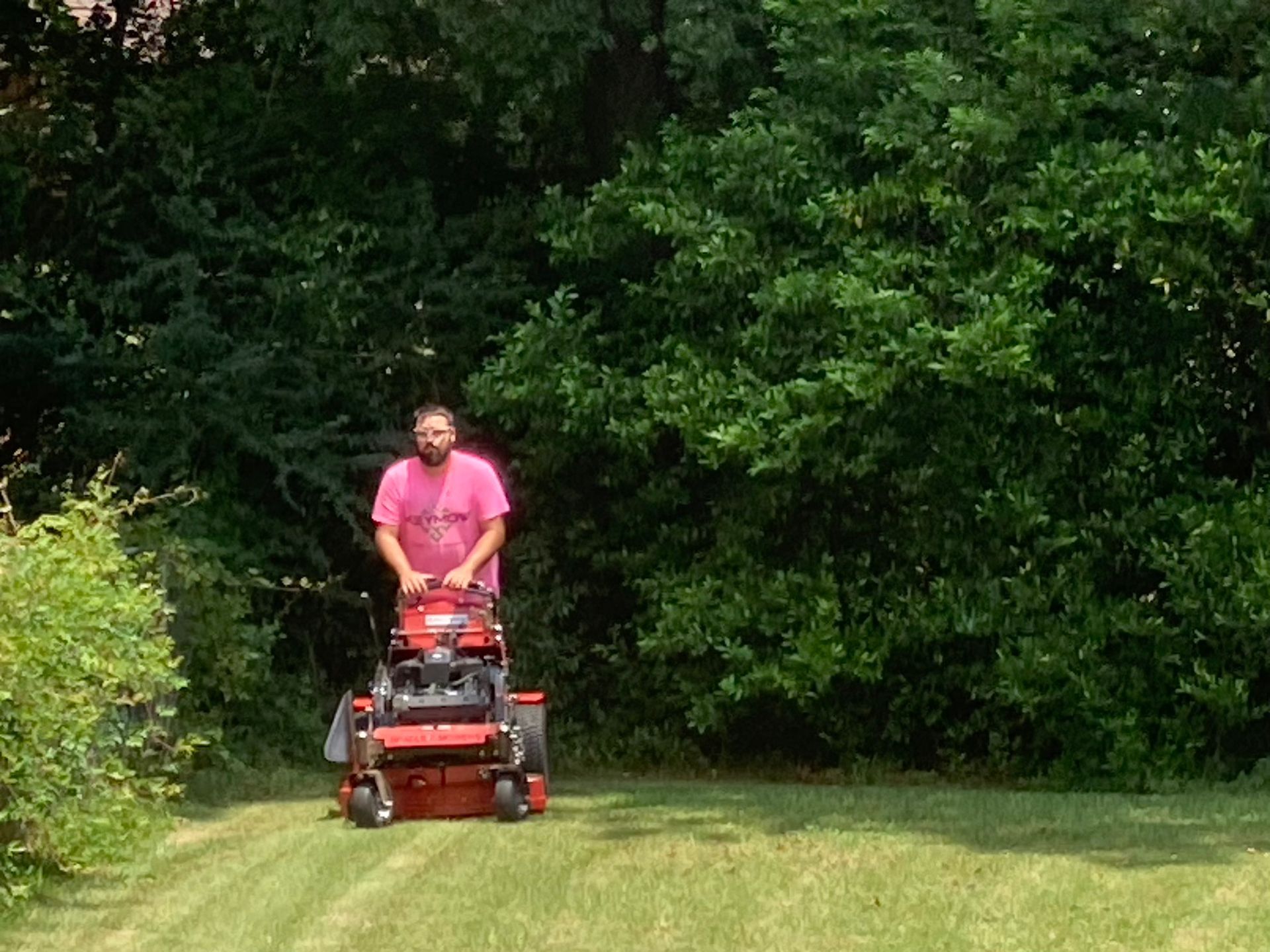 Person riding a red lawn mower on a grassy yard beside dense green trees.