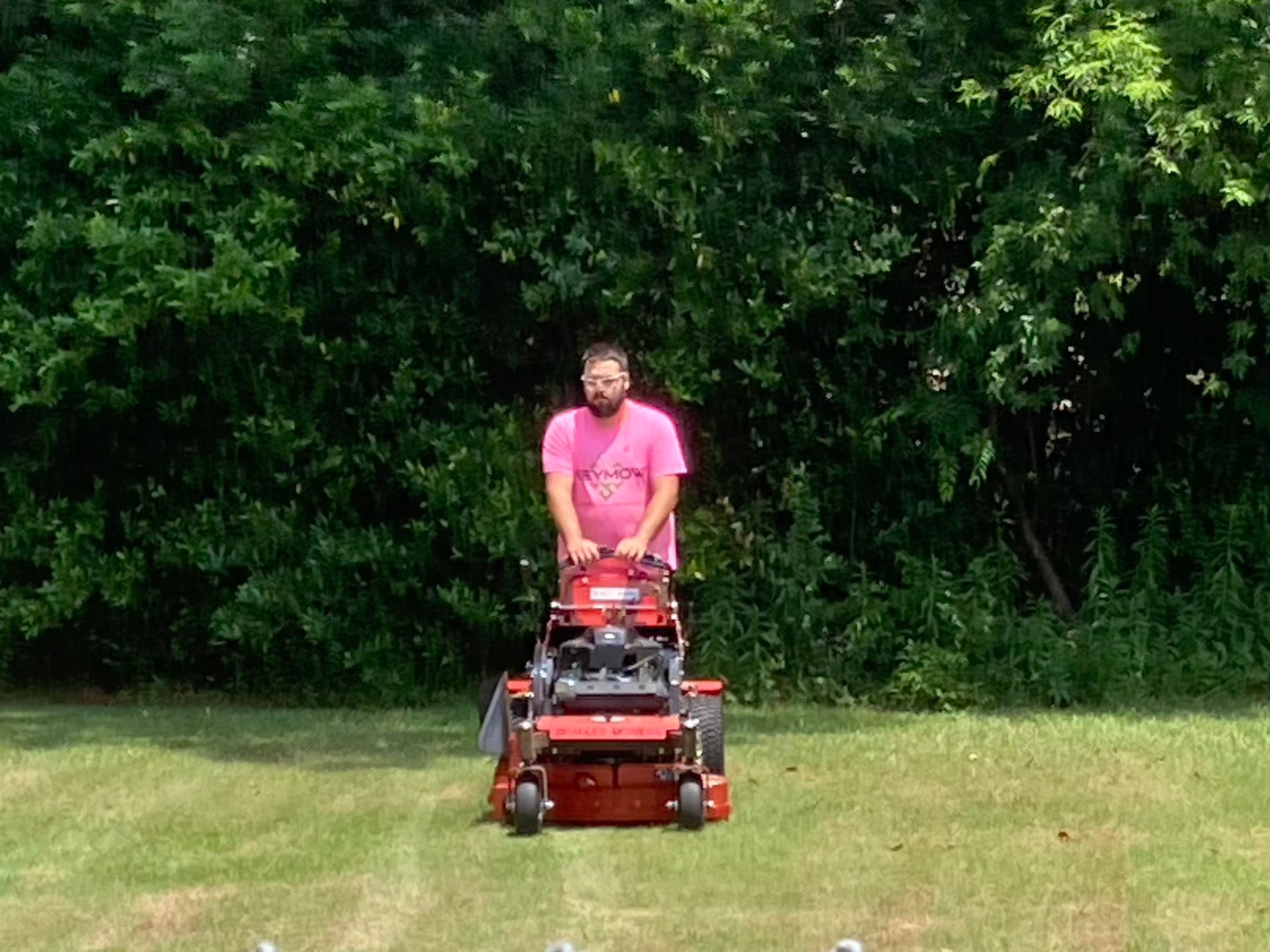 Person in a pink shirt riding a red lawn mower on grass with trees in the background