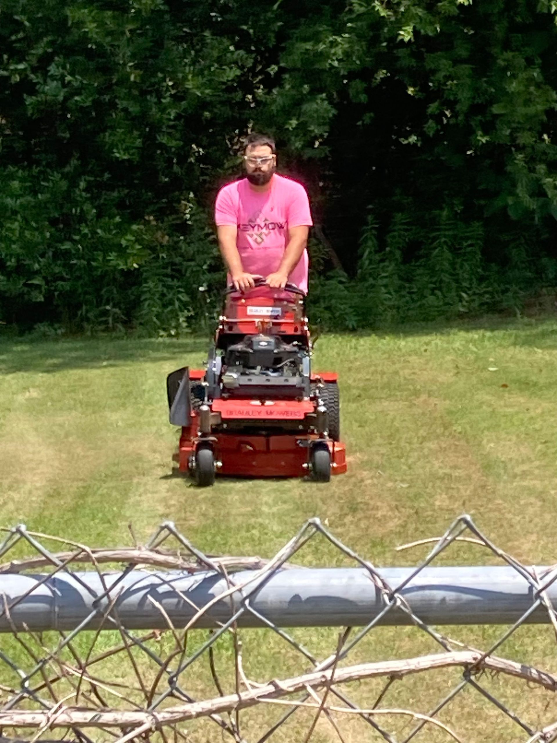 Person mowing a grassy yard with a red lawn mower, viewed through a chain-link fence.