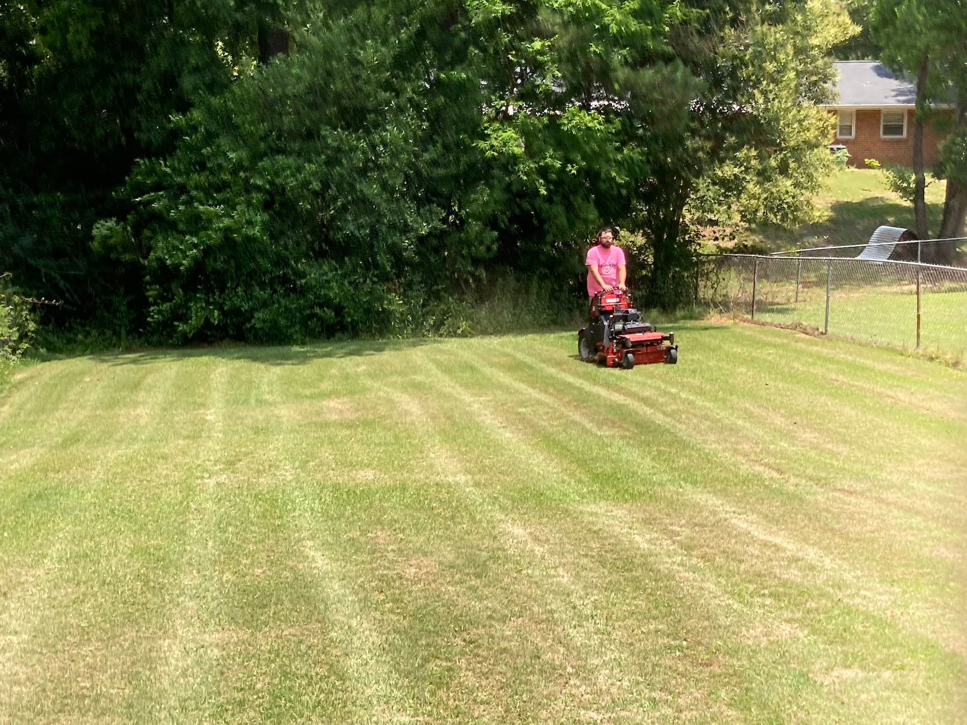 Person mowing a grassy yard with a red riding mower near trees and a fence.