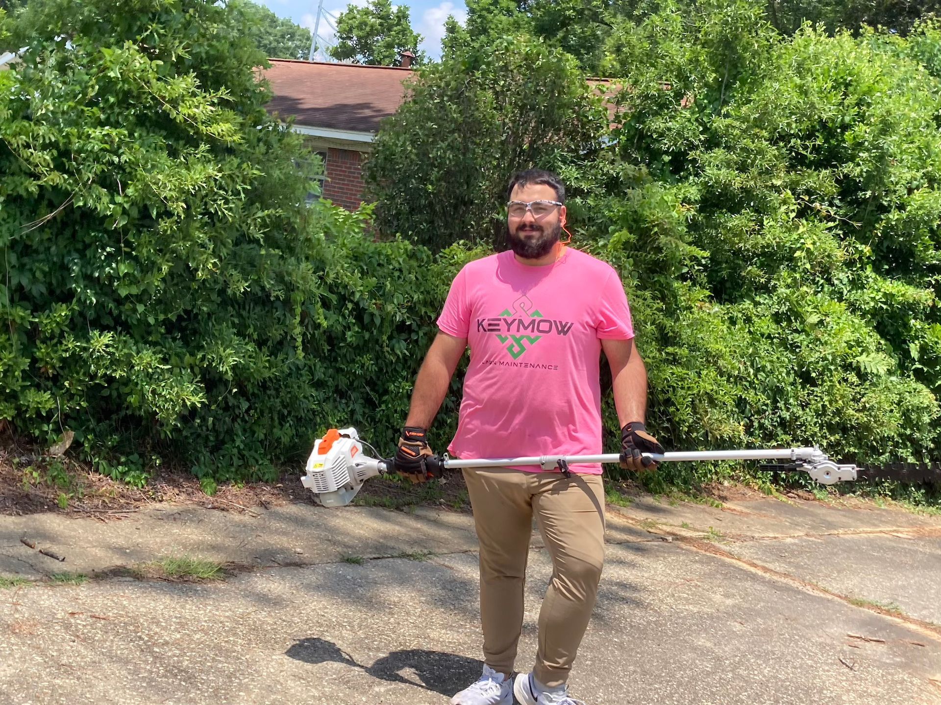 Man smiling in a pink shirt holding a string trimmer on a driveway, with green bushes behind him