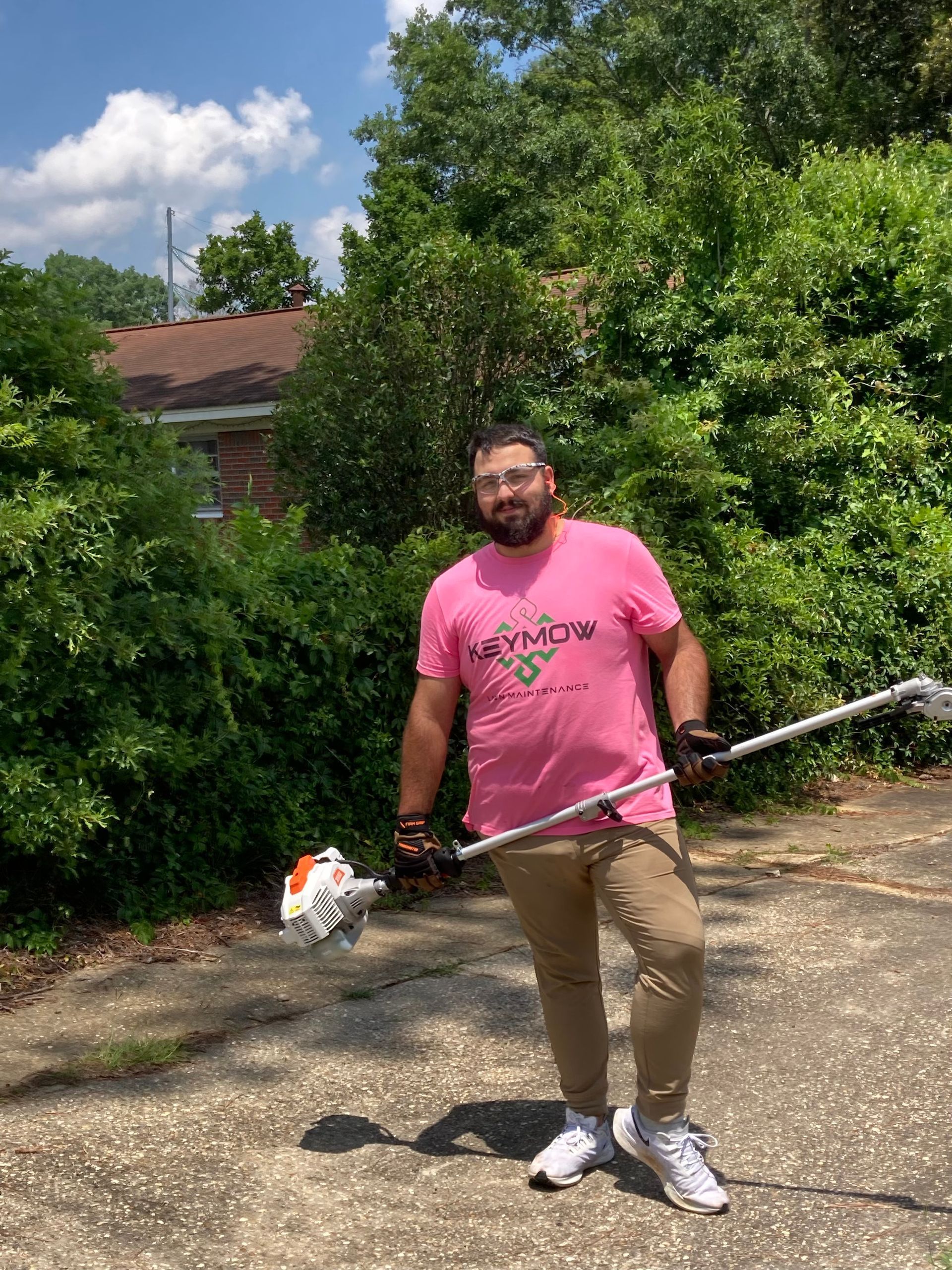 Person in a pink shirt holding a long pole outdoors on a sunny driveway, with trees and a house behind.