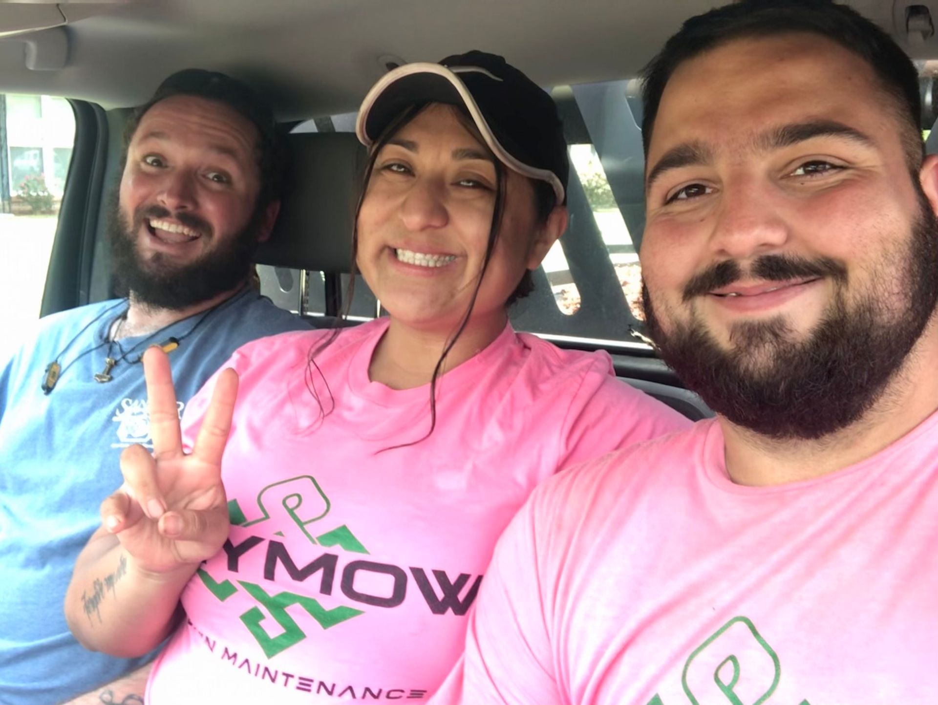 Three smiling people in a car wearing matching pink shirts, one making a peace sign