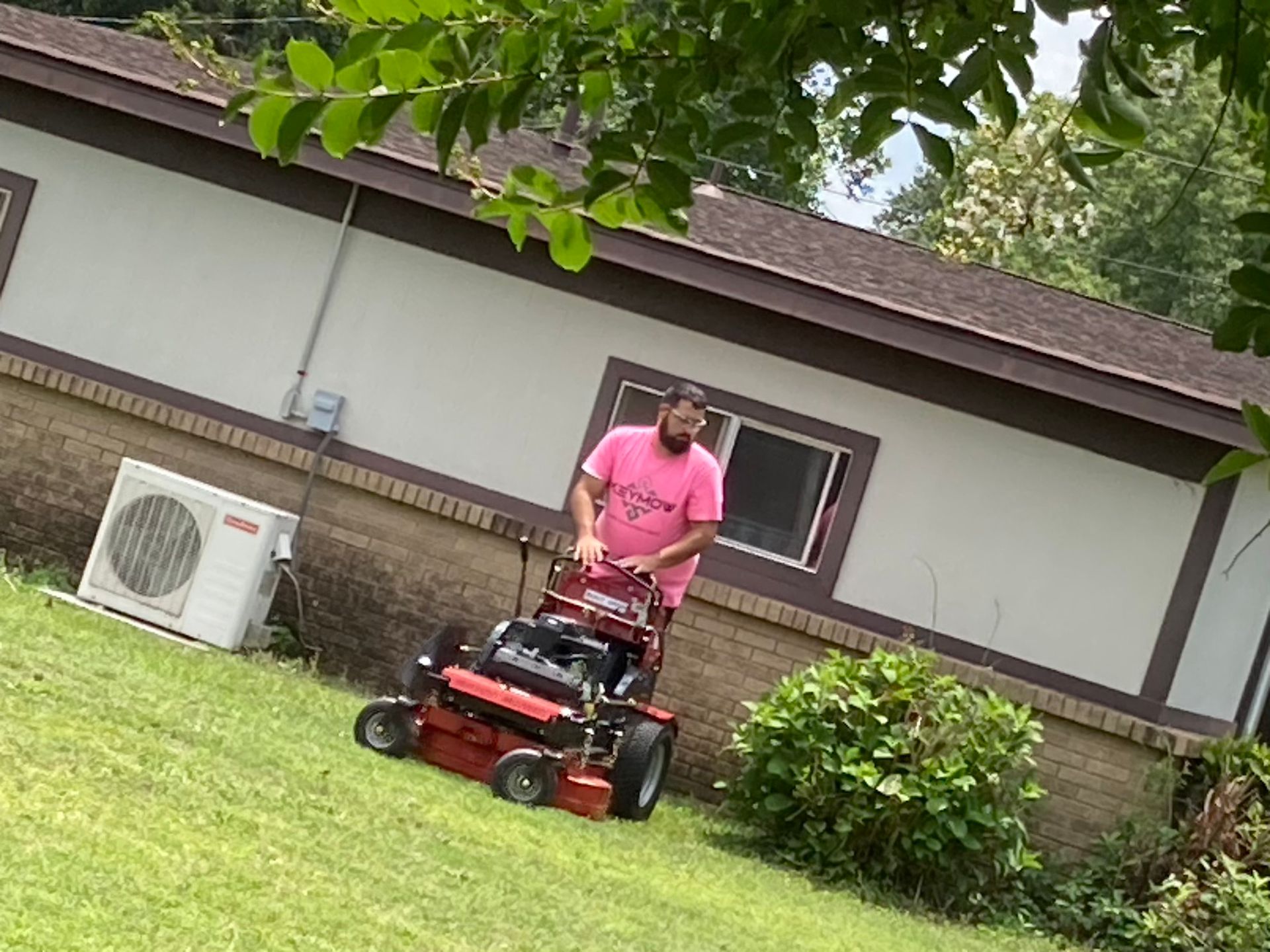 Person mowing a lawn with a red riding mower beside a house and shrubs.