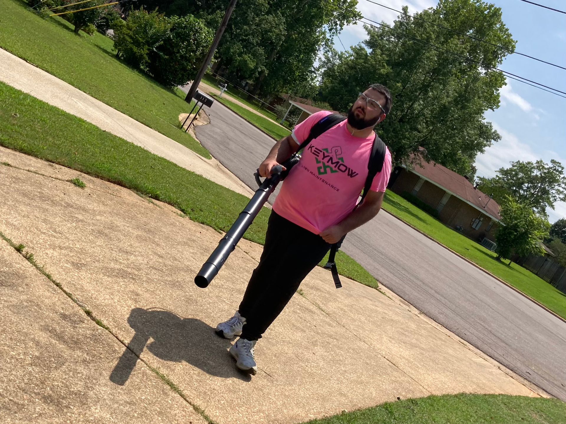 Person in a pink shirt stands on a driveway holding a tripod, with a suburban street and lawn behind.