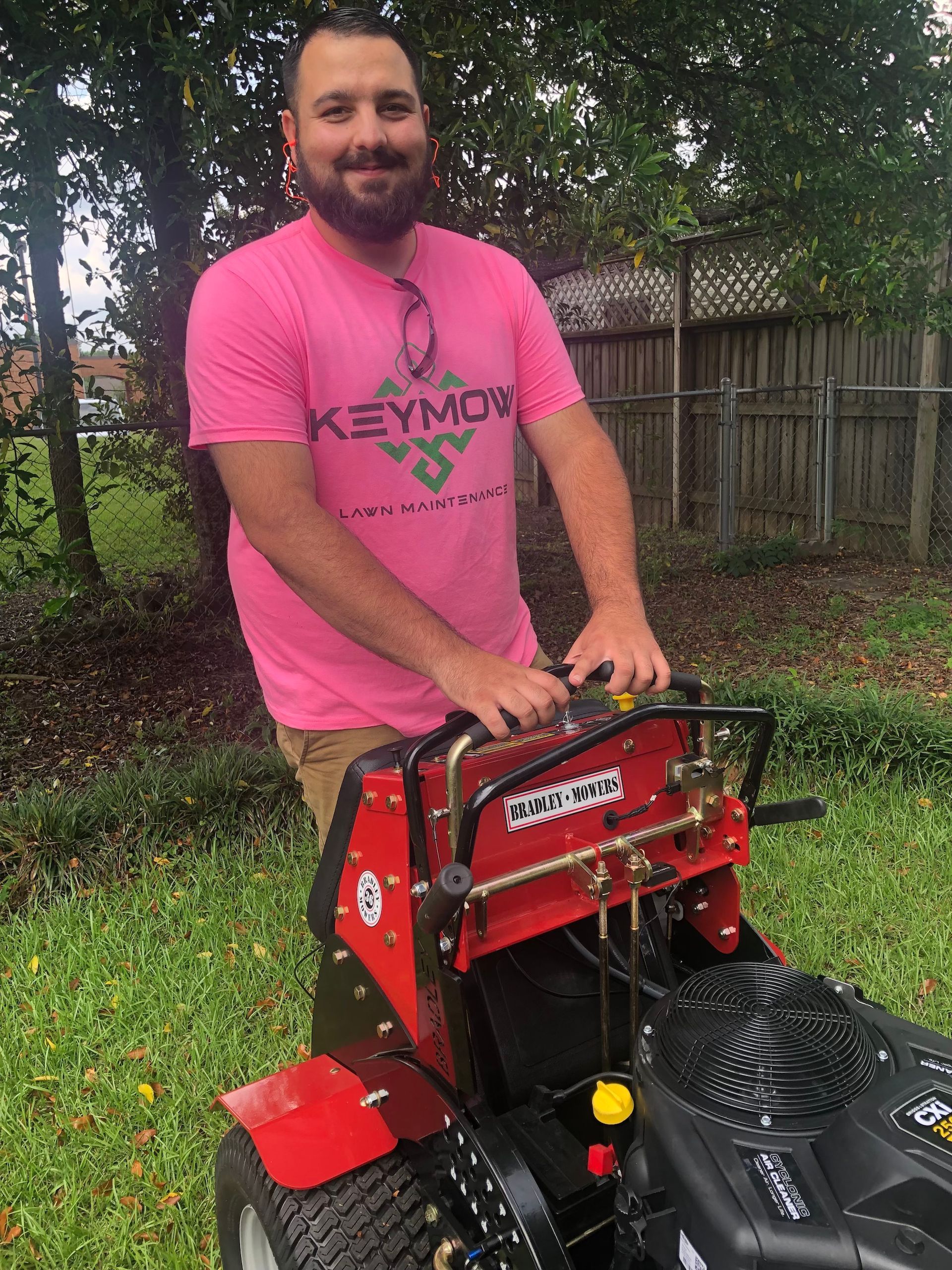 Man in pink shirt standing beside a red lawn machine in a grassy backyard