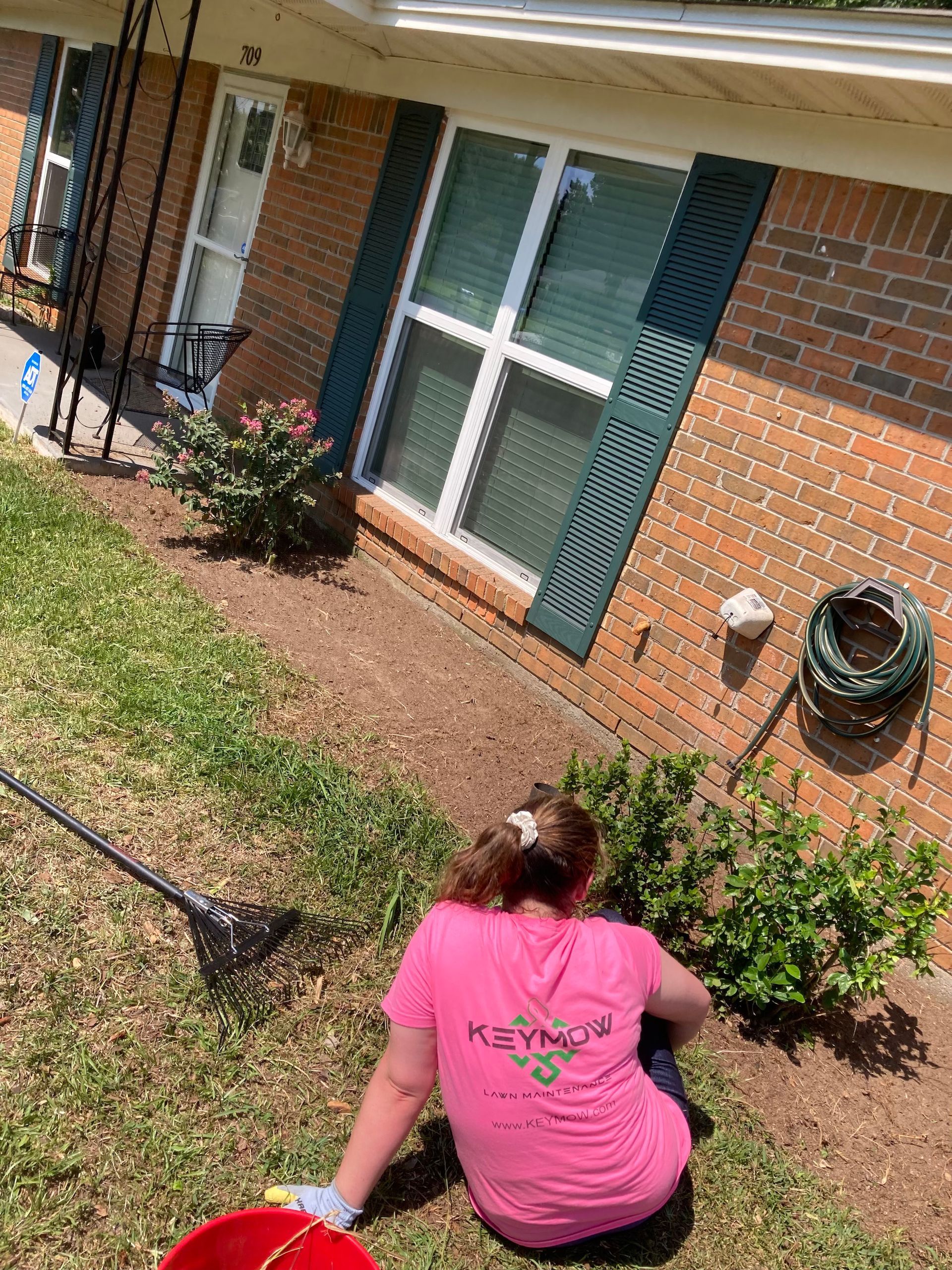 Person kneeling in a pink shirt in a yard, trimming or pulling weeds beside a brick house window.