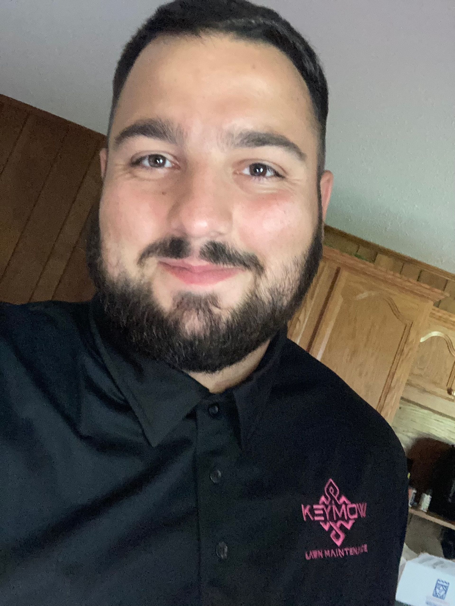 Smiling man in a black polo shirt standing indoors in front of wooden cabinets.