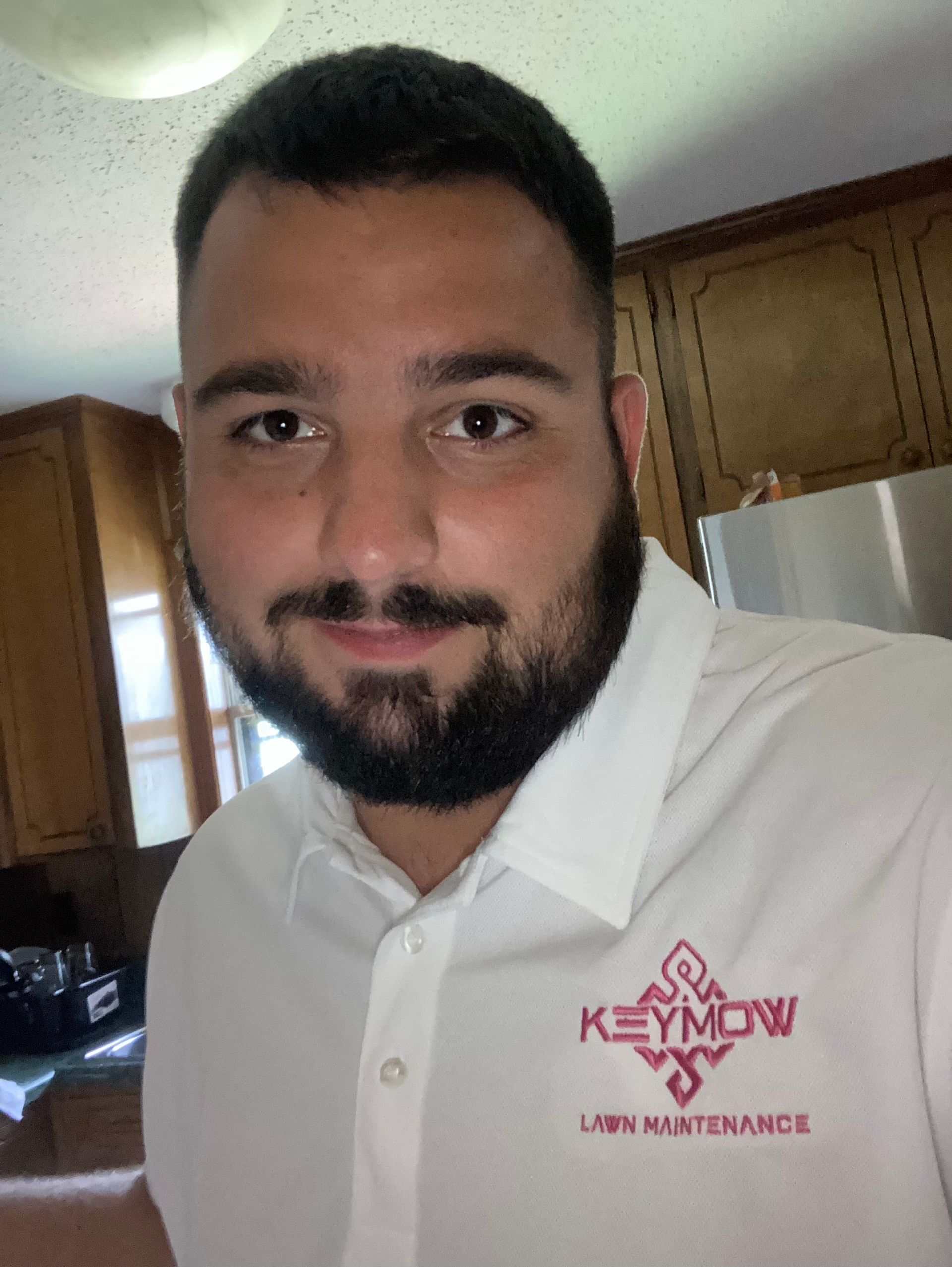 Man in a white polo shirt with a red logo, smiling indoors in a kitchen.