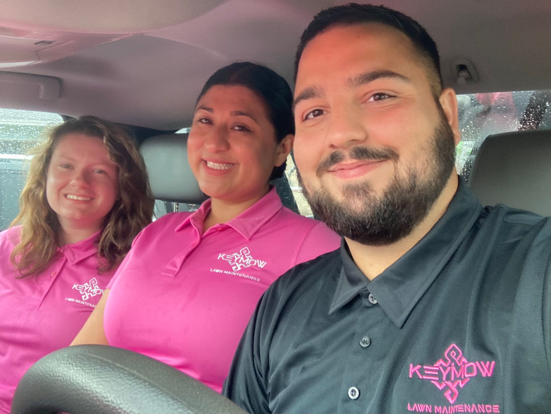 Three people in pink and black polos smiling inside a car
