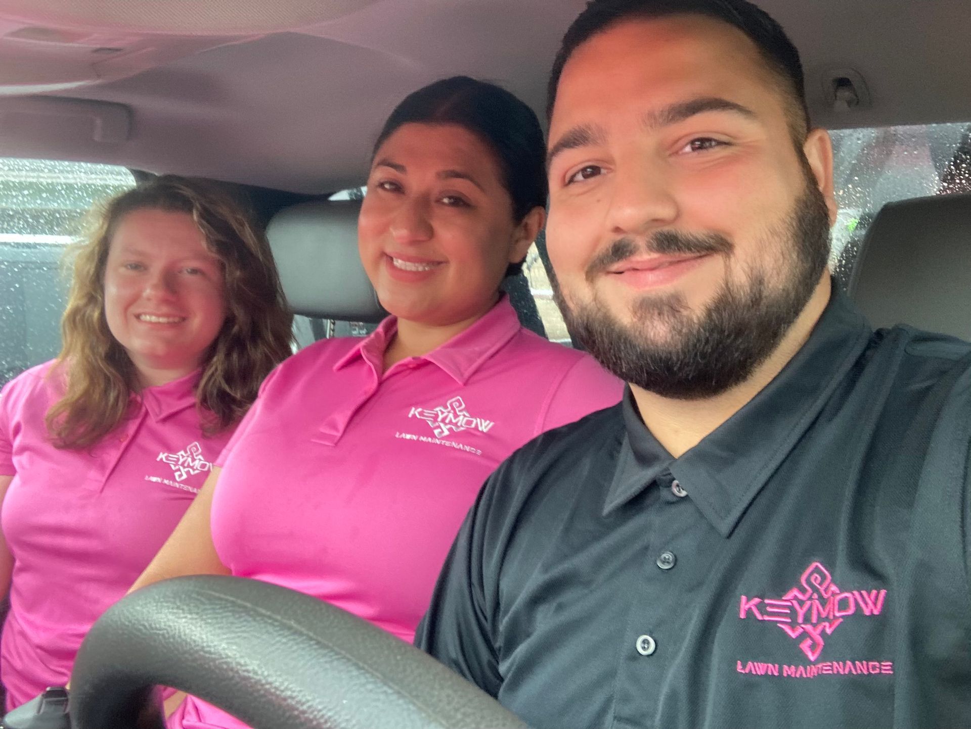 Three staff members in pink shirts smiling inside a car