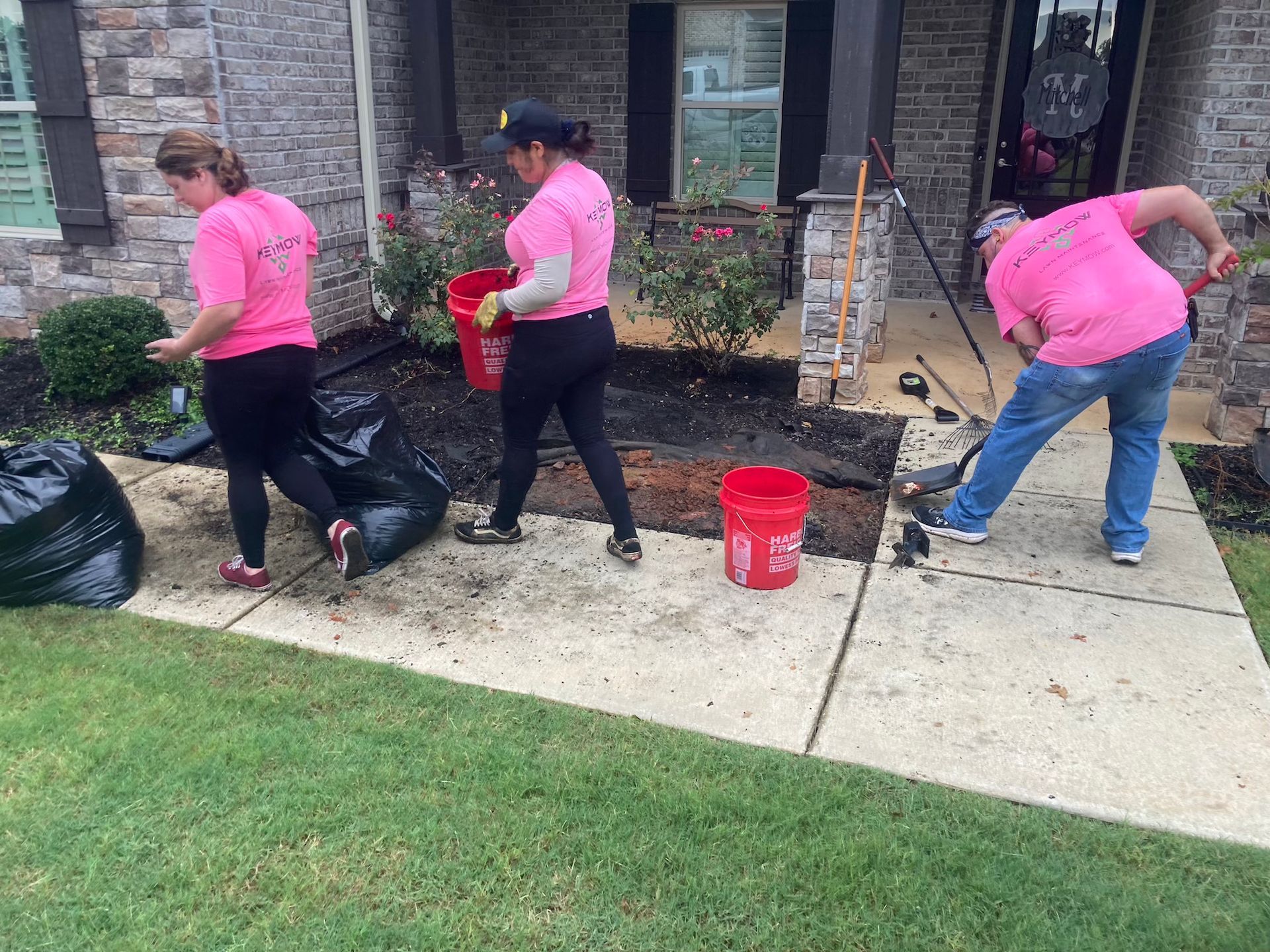 Three volunteers in pink shirts bag leaves and debris outside a house.