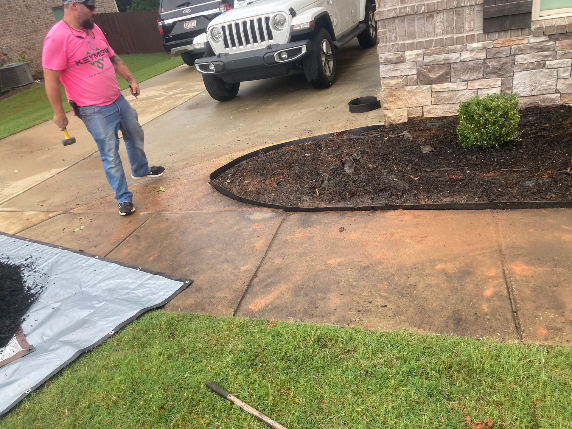 Worker in pink shirt on a driveway near a parked Jeep and a stone house facade.
