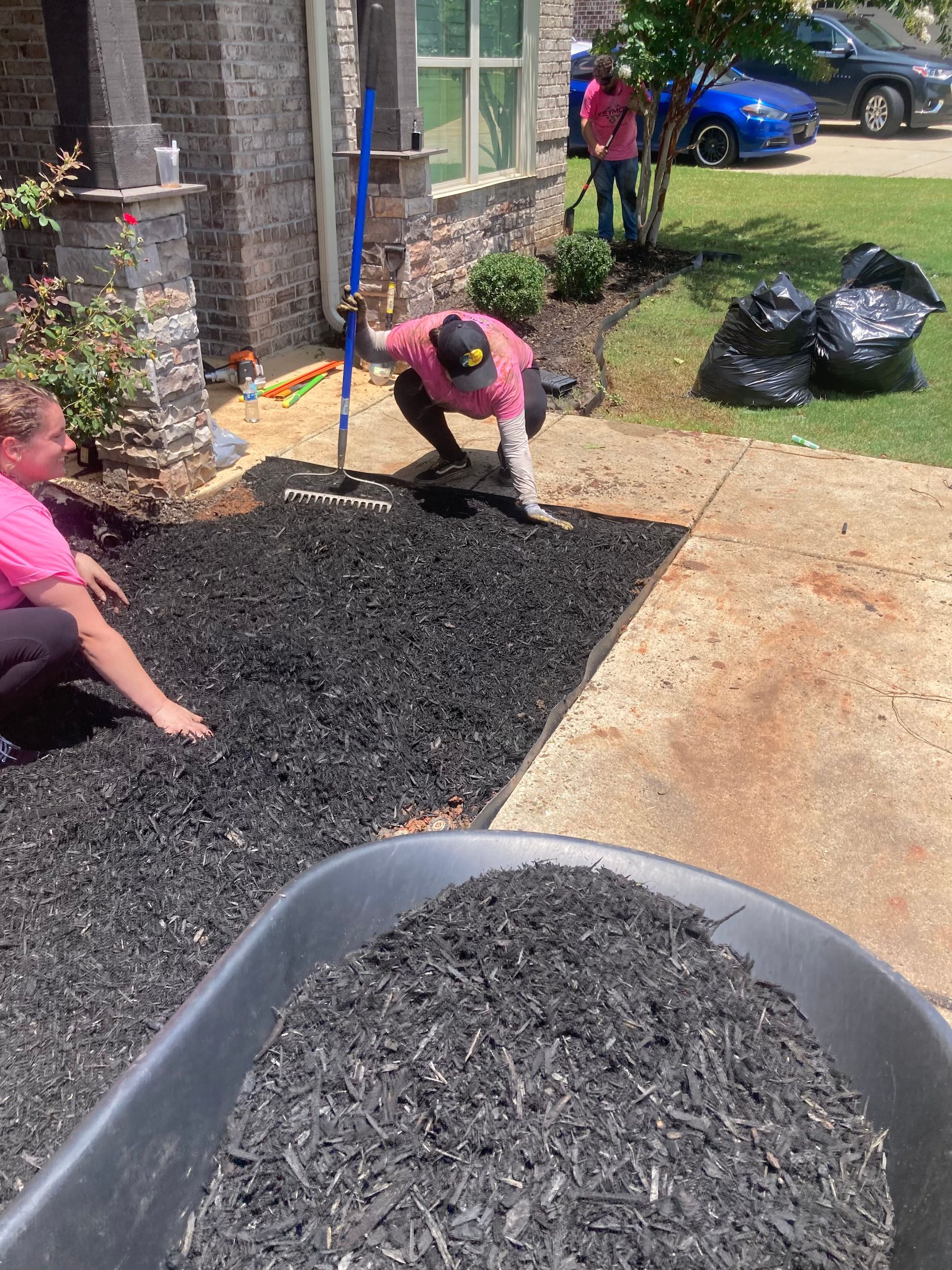 People spreading black mulch beside a brick house and driveway, with trash bags nearby.