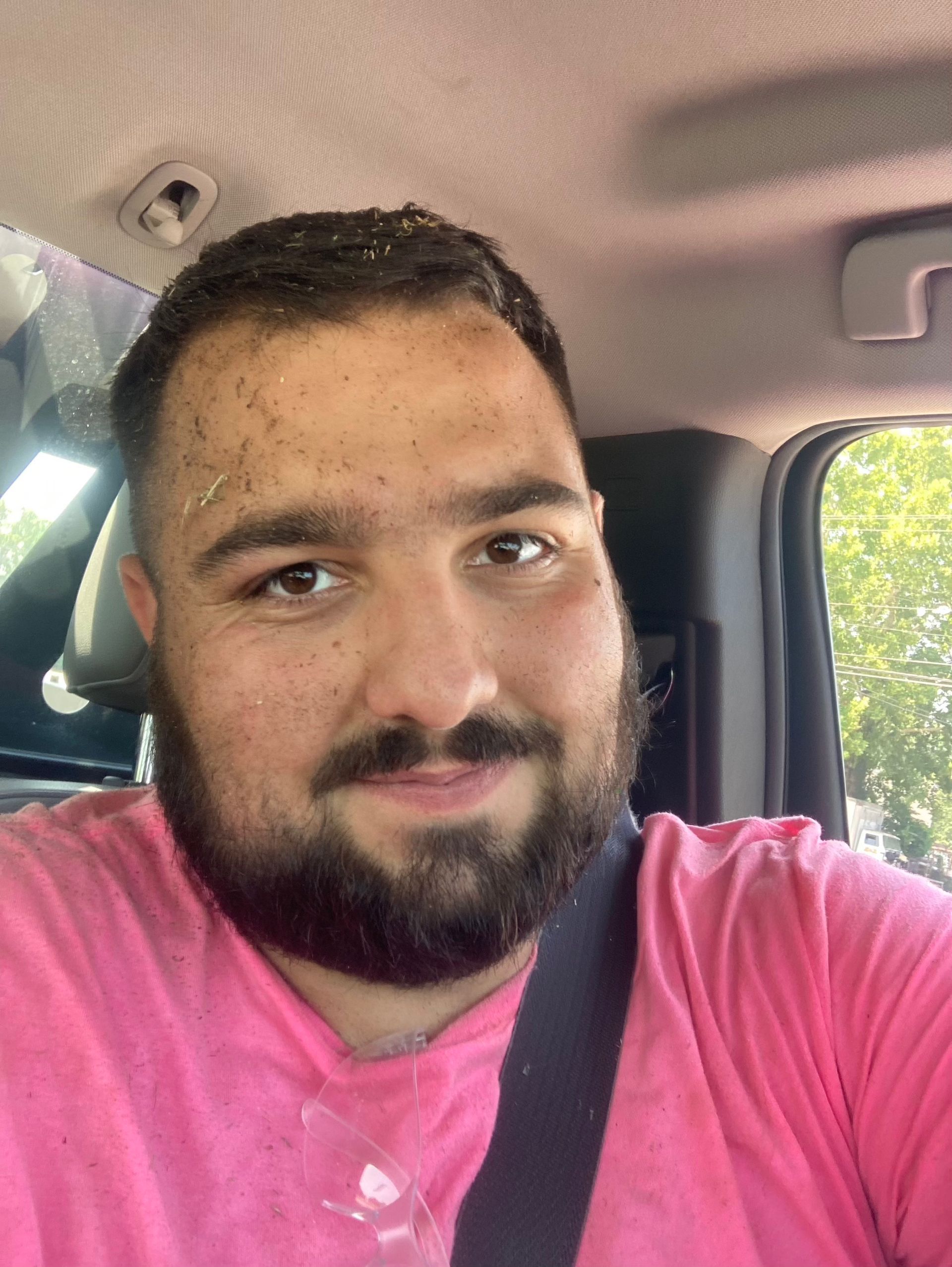 Person with short dark hair and beard in a pink shirt sitting inside a car, smiling at the camera.