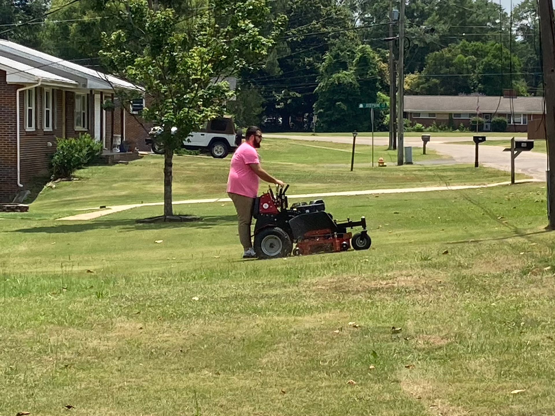 Person mowing a grassy lawn with a riding mower beside a brick building and trees