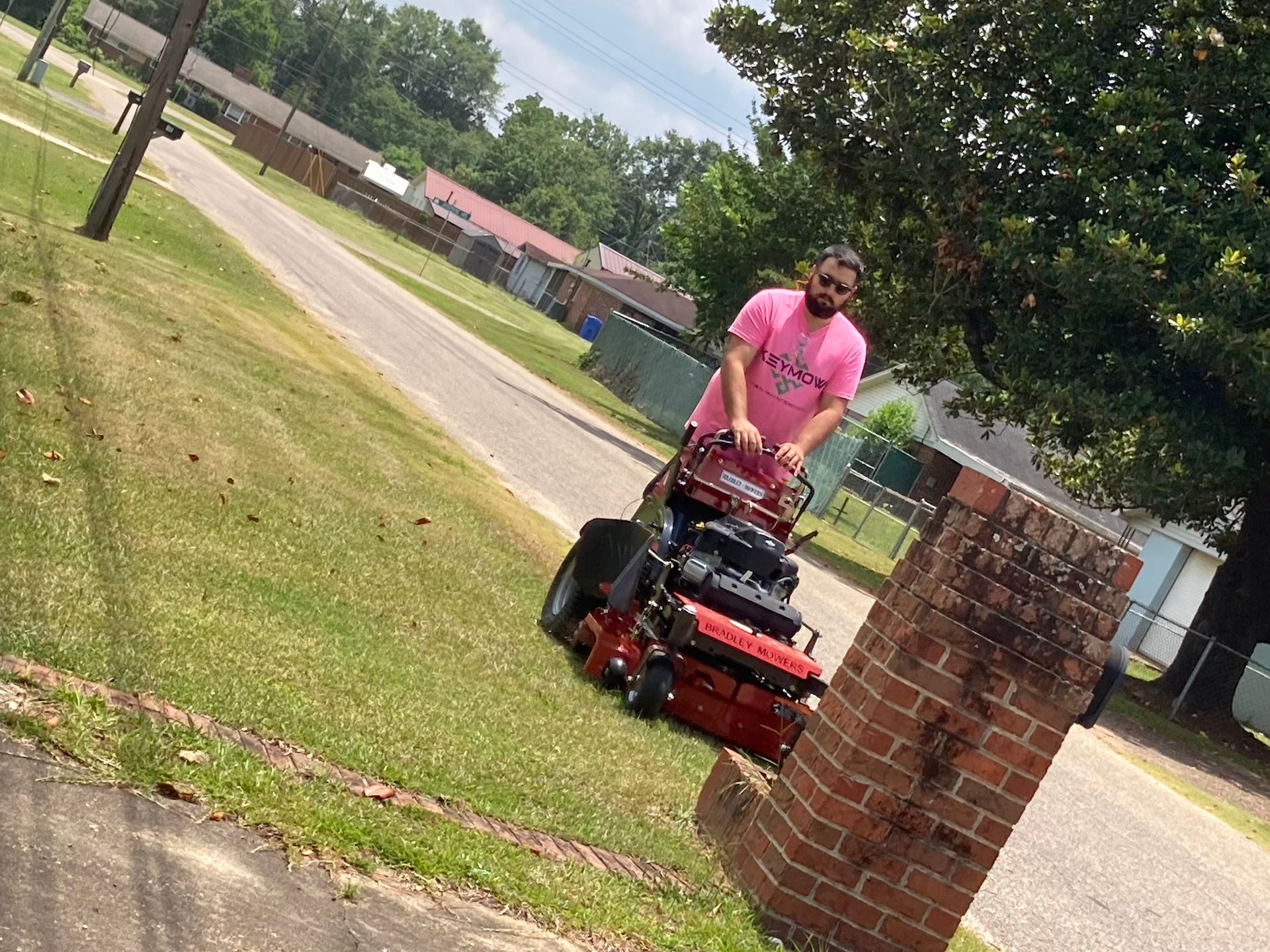 Person mowing a lawn with a red riding mower near a brick mailbox in a suburban yard