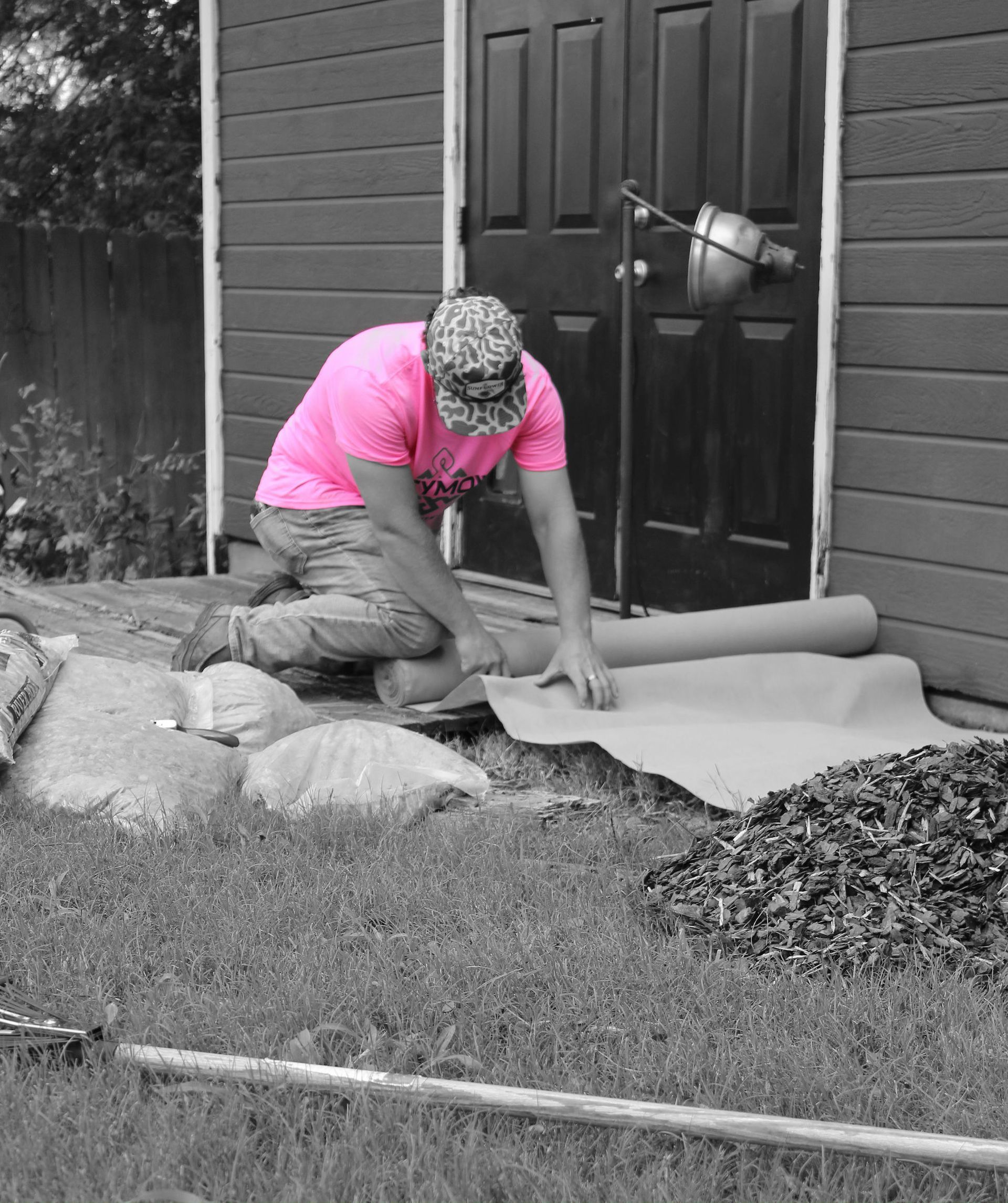 Person kneeling on a mat by a shed door, rolling out material on the ground in a backyard.