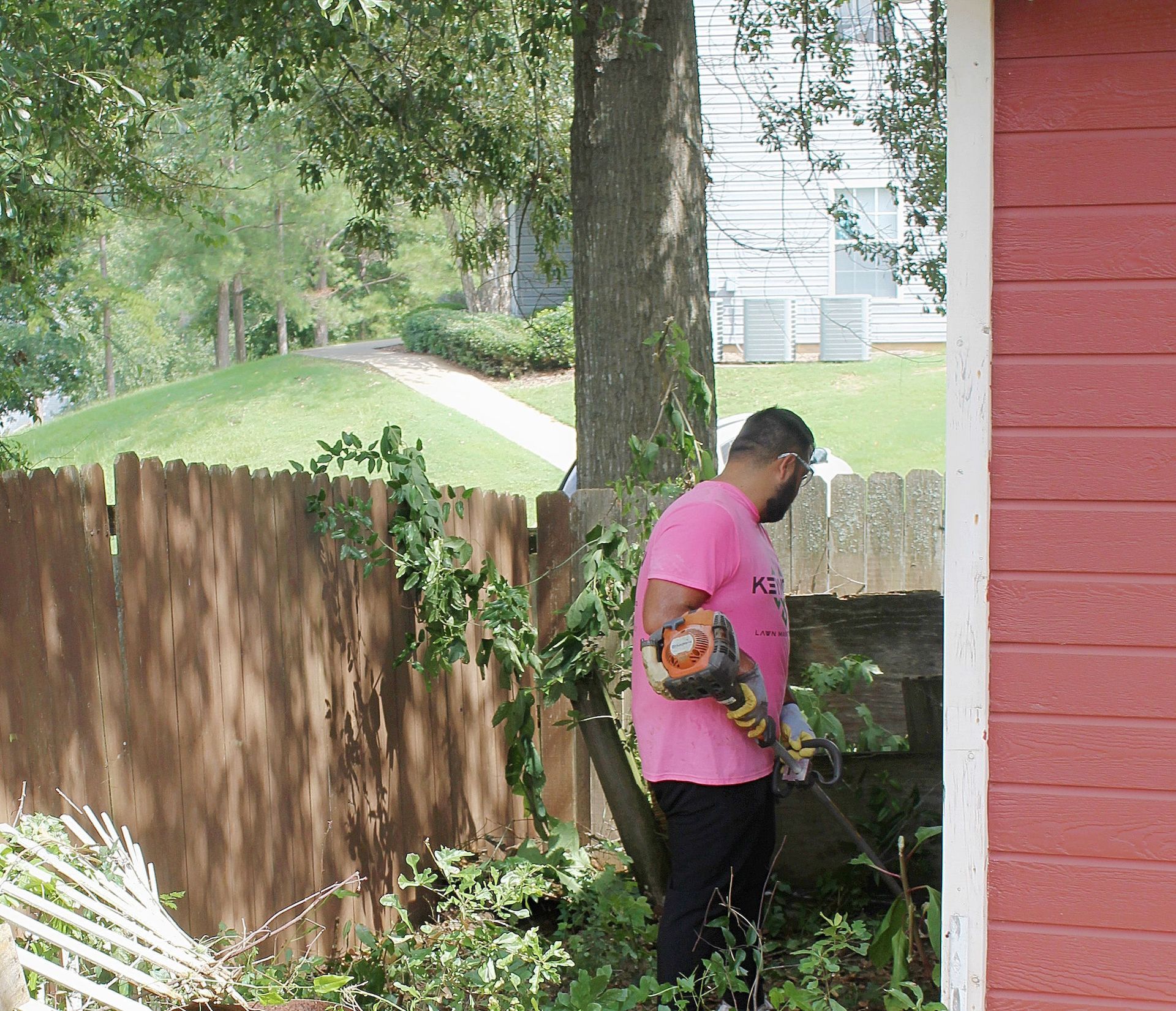 Person using a chainsaw beside a red house in a backyard, near a tree and wooden fence.