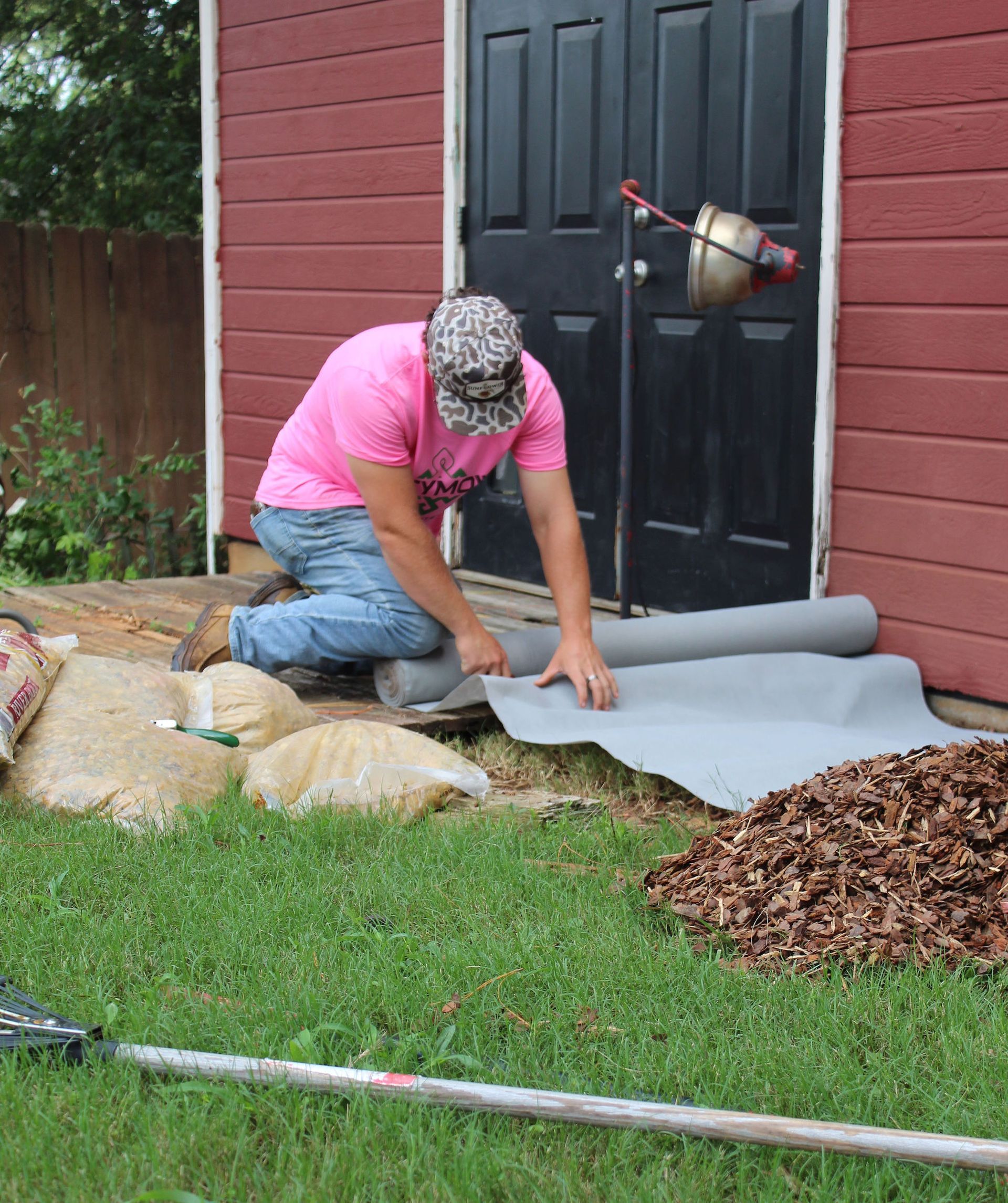 Person rolling gray underlayment on a backyard lawn beside a house and patio door