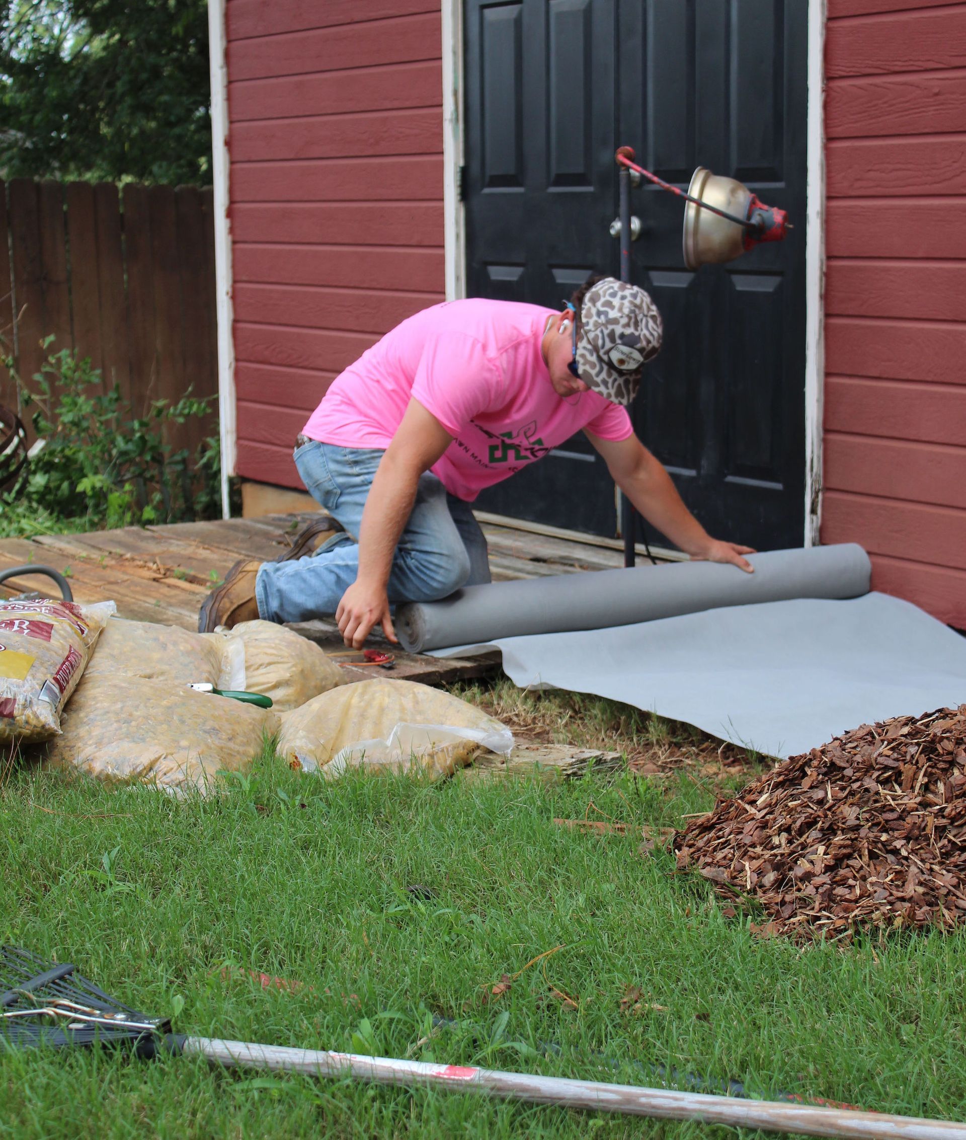 Person in a pink shirt rolling out gray fabric beside a shed in a yard