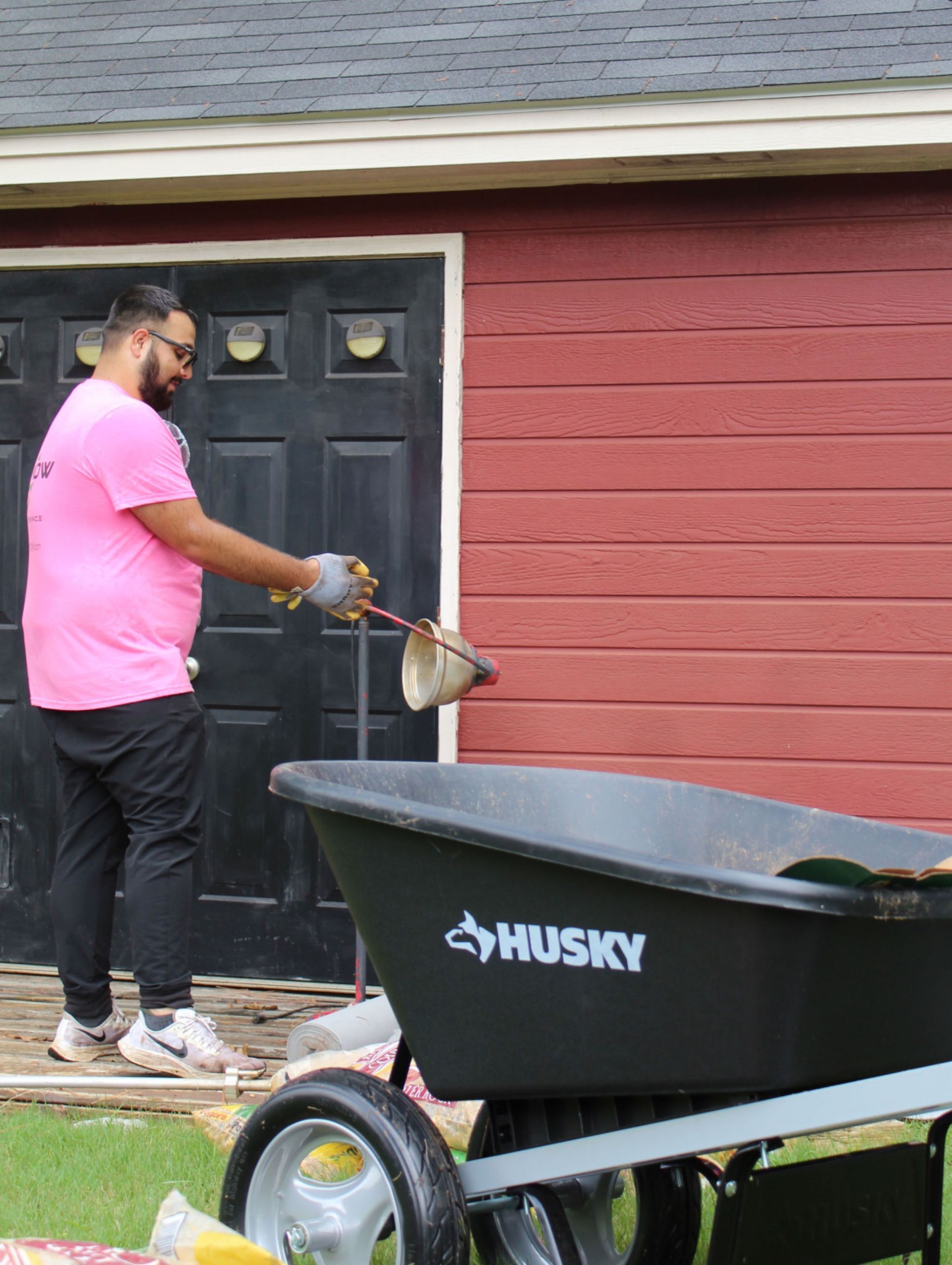 Person spraying a red house wall near a black door beside a Husky wheelbarrow