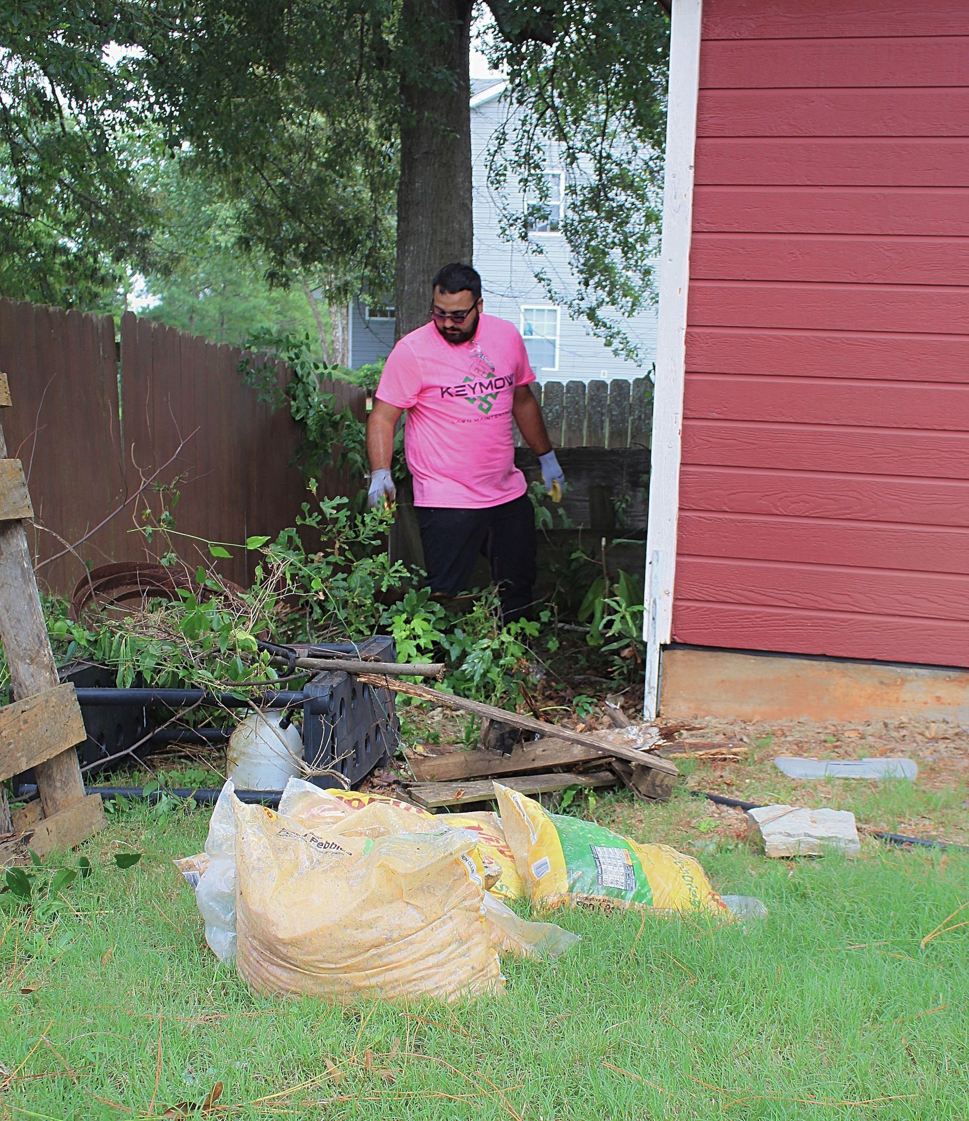 Person in pink shirt standing in a backyard beside yard debris and a red house.