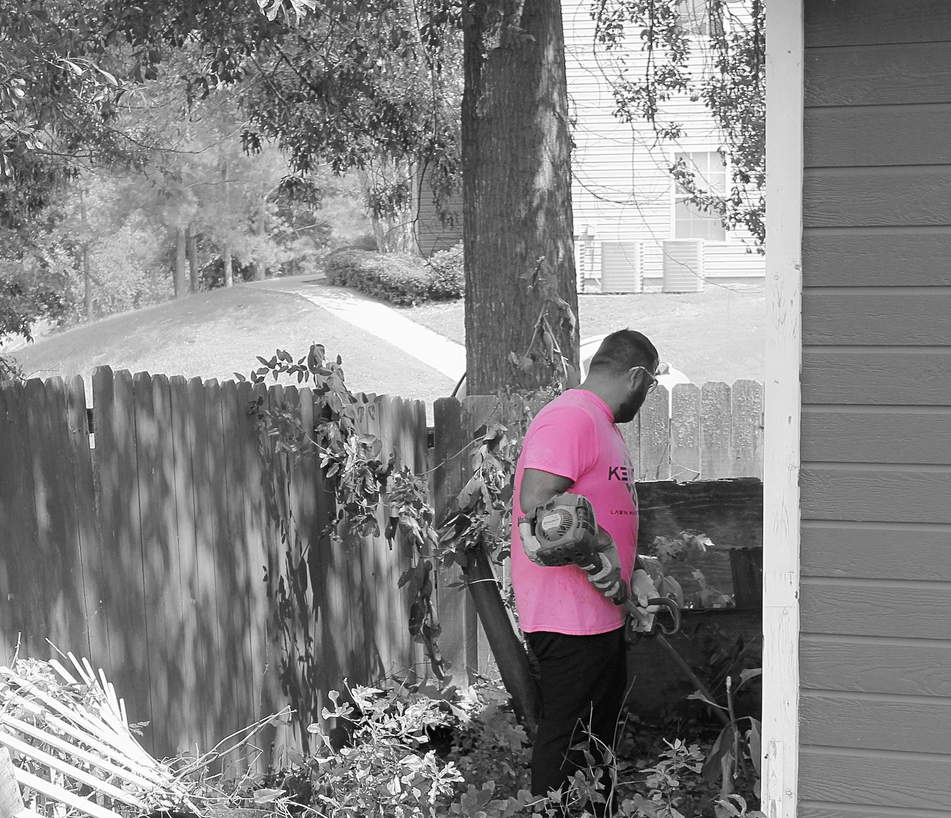 Person in a bright pink shirt works beside a tree in a backyard near a fence and house.