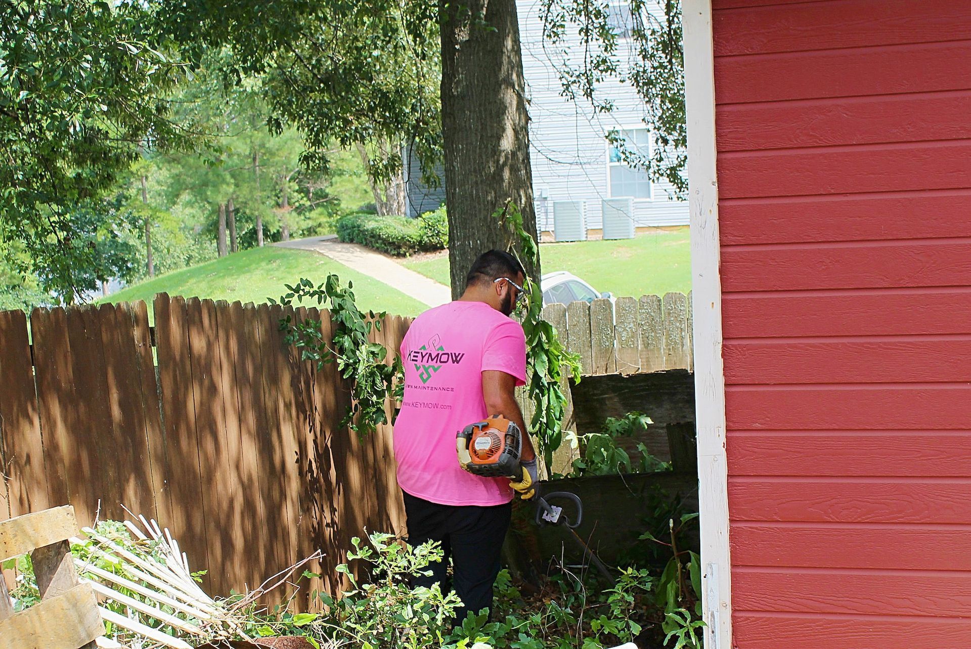Person in a pink shirt gardening beside a red house, with trees and a wooden fence nearby.