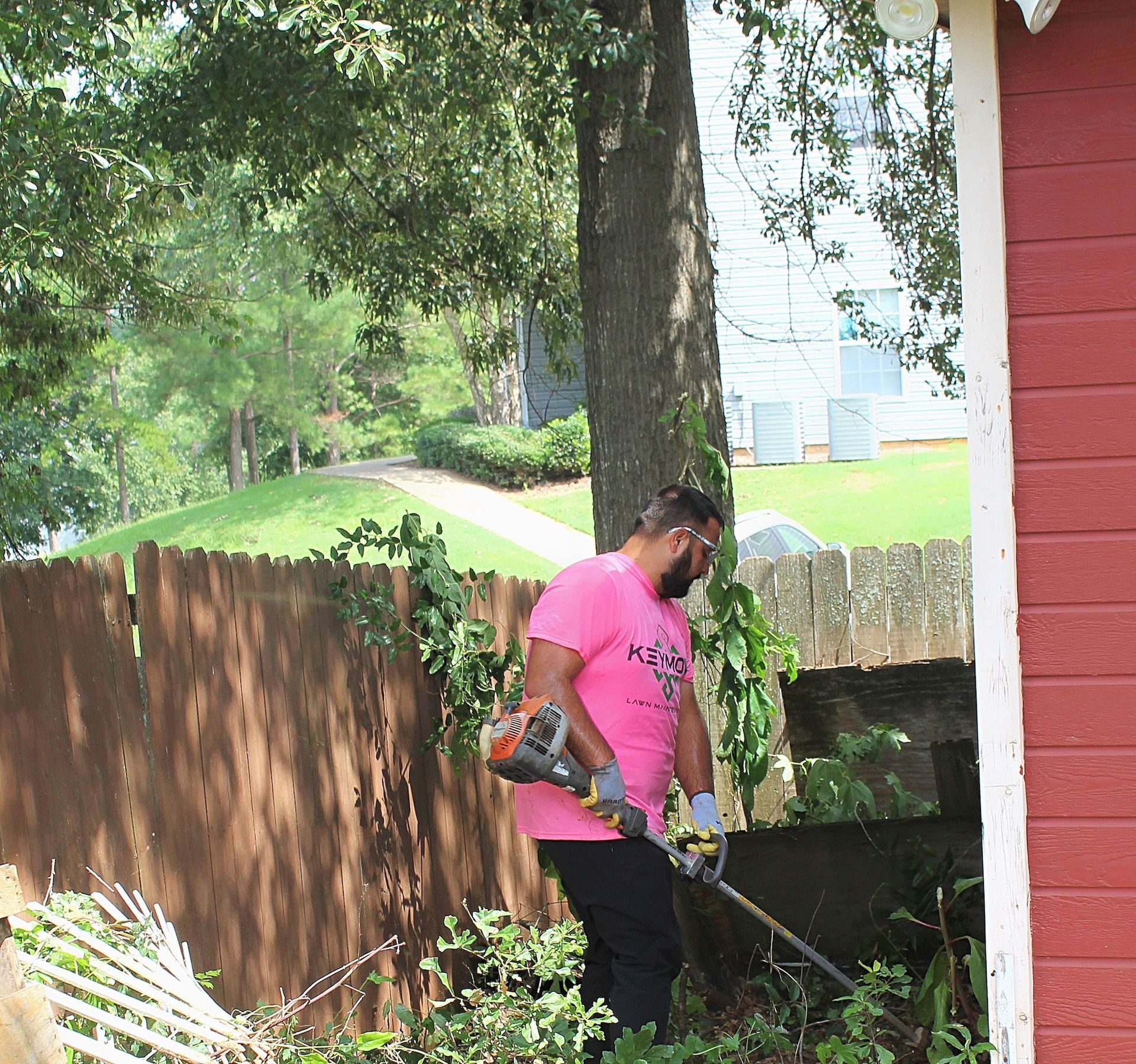 Person in a pink shirt using a string trimmer beside a red house and wooden fence.