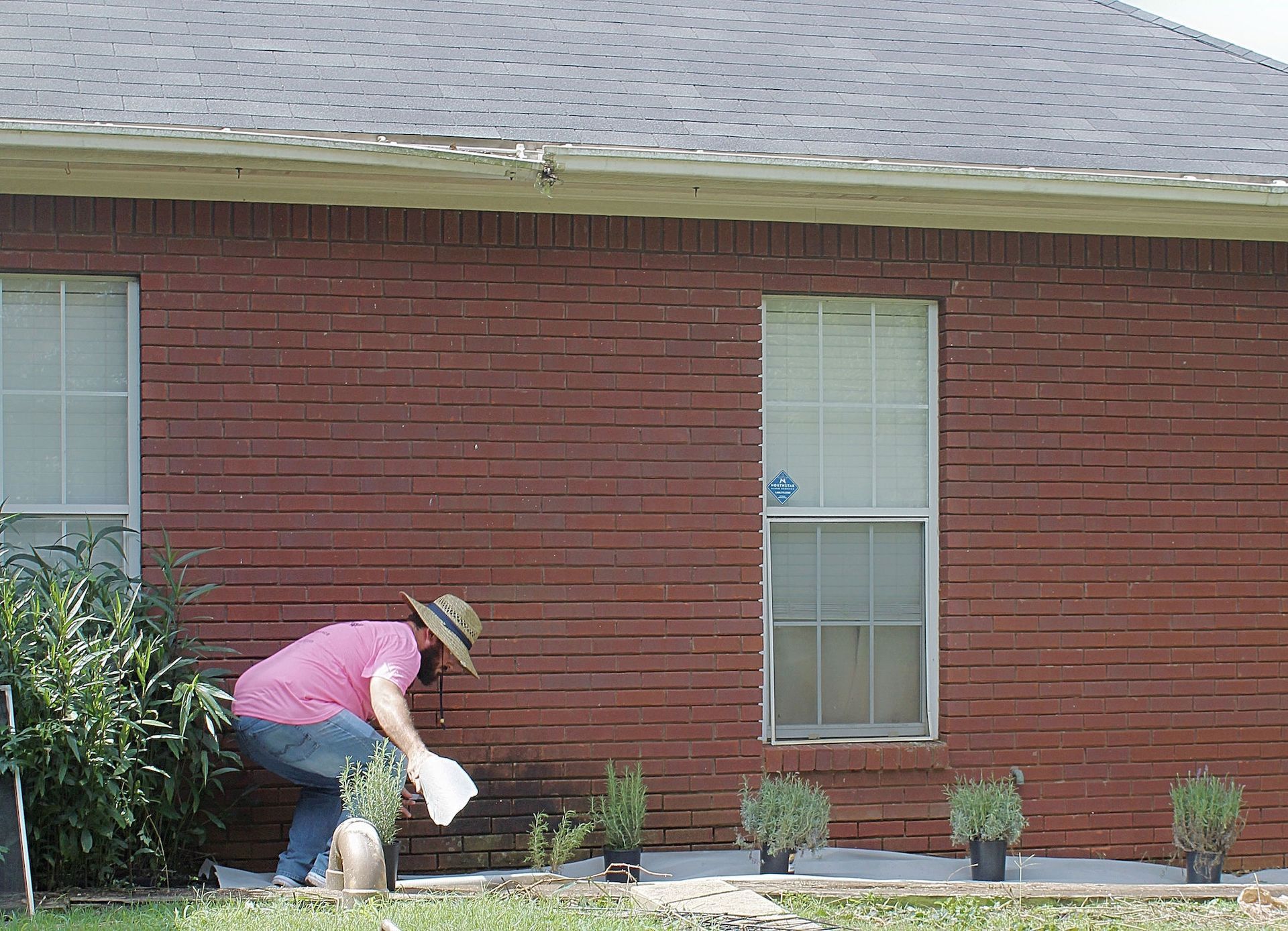 Person kneels in a front yard planting flowers beside a red brick house.