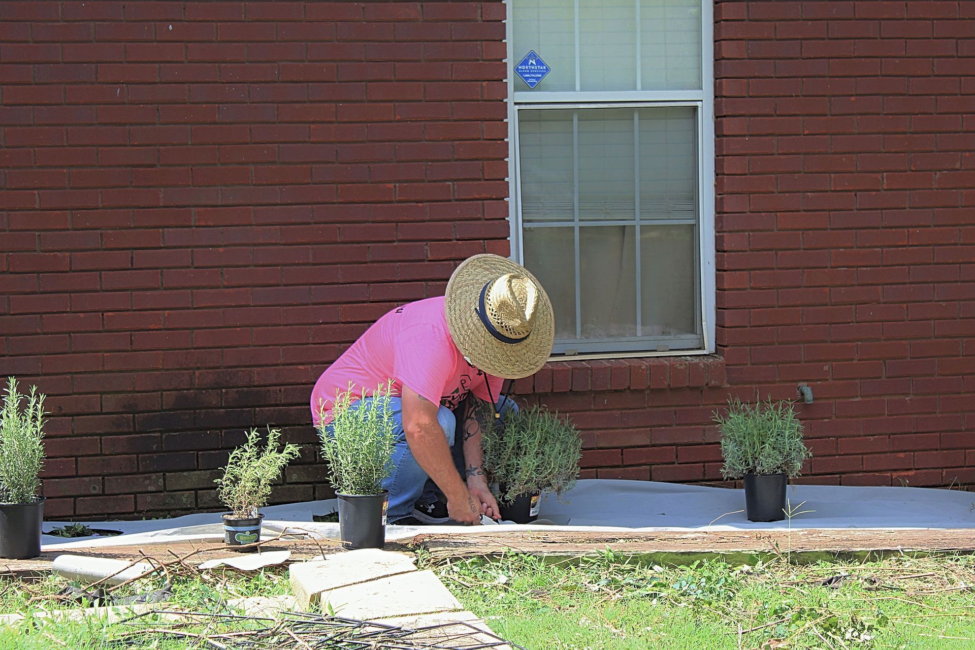 Person in a pink shirt and straw hat kneeling by potted plants beside a brick house.