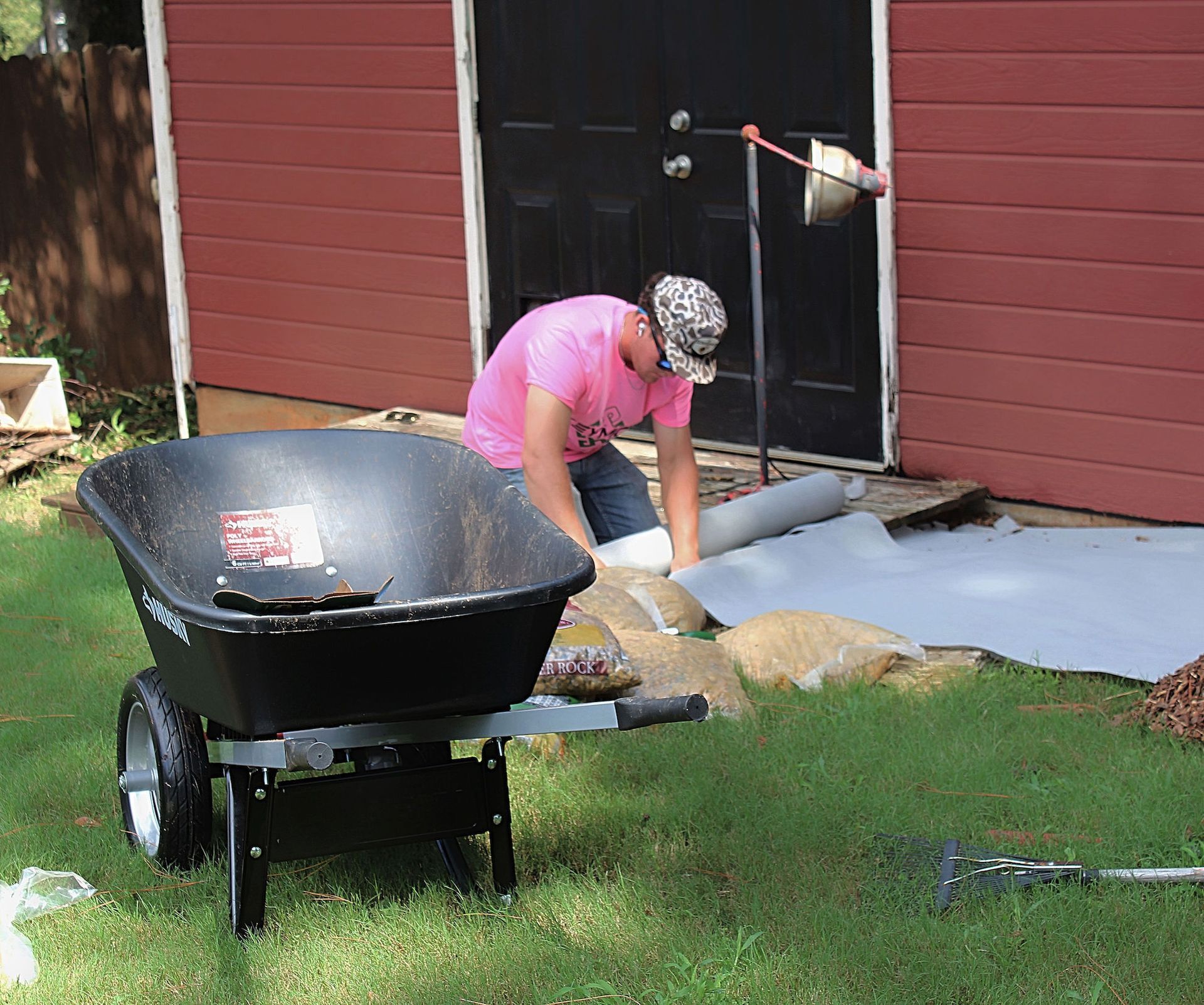 Person in pink shirt working in a yard beside a black wheelbarrow near a red house.