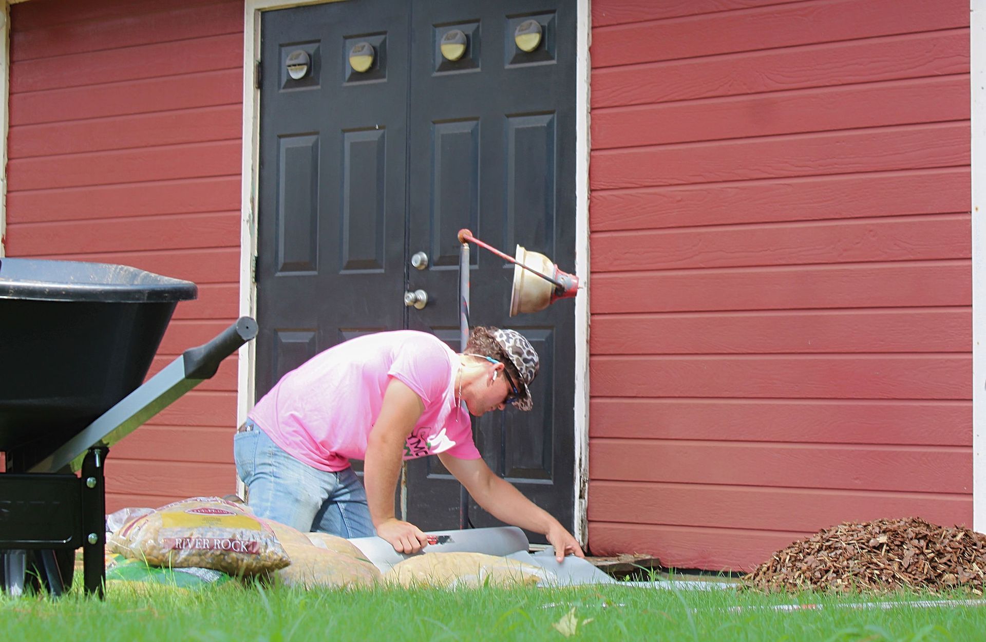 Person kneeling by a black door, planting near a red house with a wheelbarrow and a bag of soil.