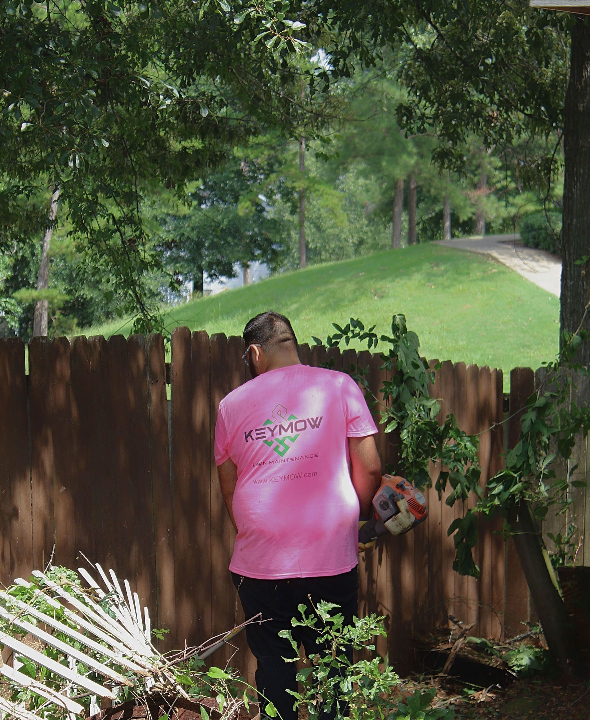 Person in a pink shirt stands by a wooden fence in a leafy backyard holding flowers.