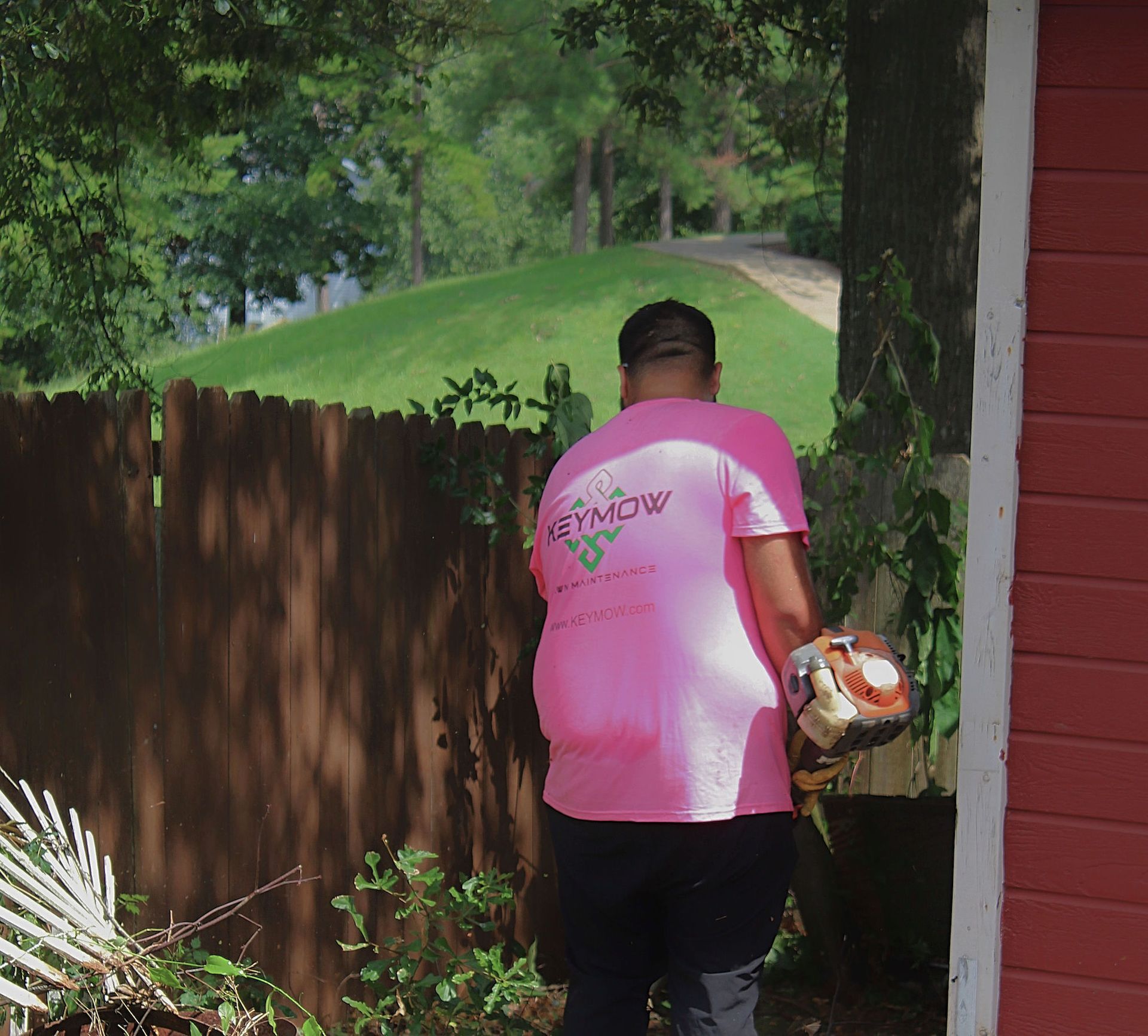 Person in pink shirt carries items beside a wooden fence and red house in a leafy backyard.