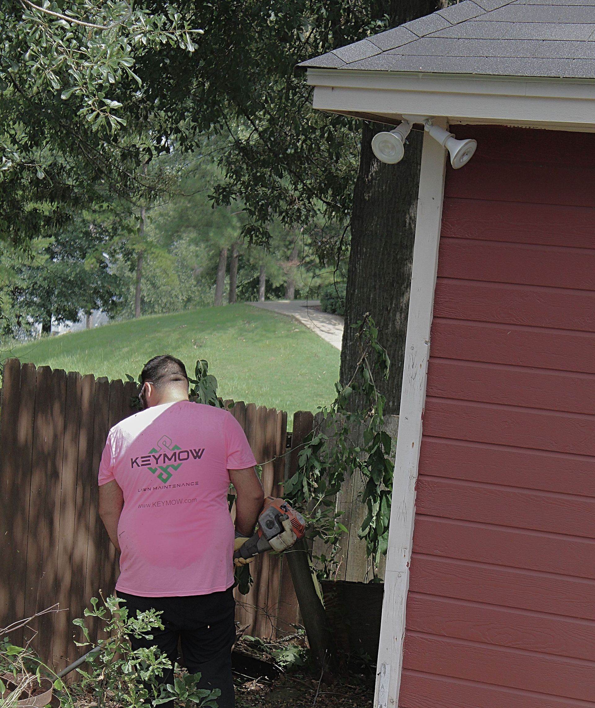 Person in pink shirt standing by a wooden fence beside a red house in a leafy yard.
