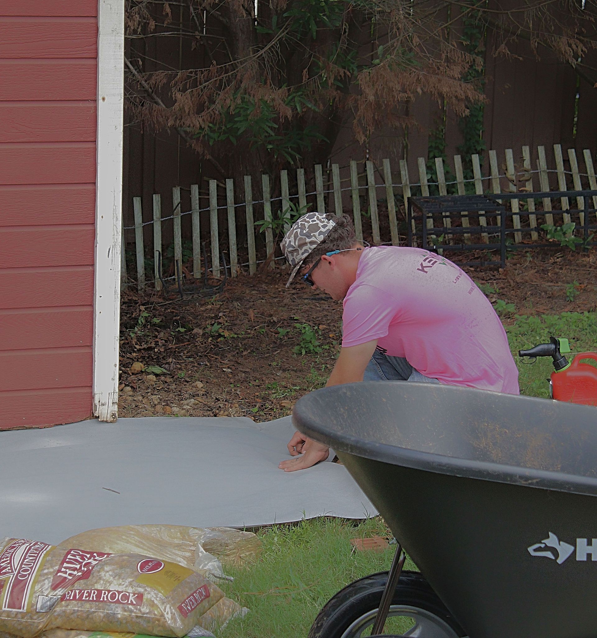Person crouching on a driveway near a red building, with a wheelbarrow in the foreground.