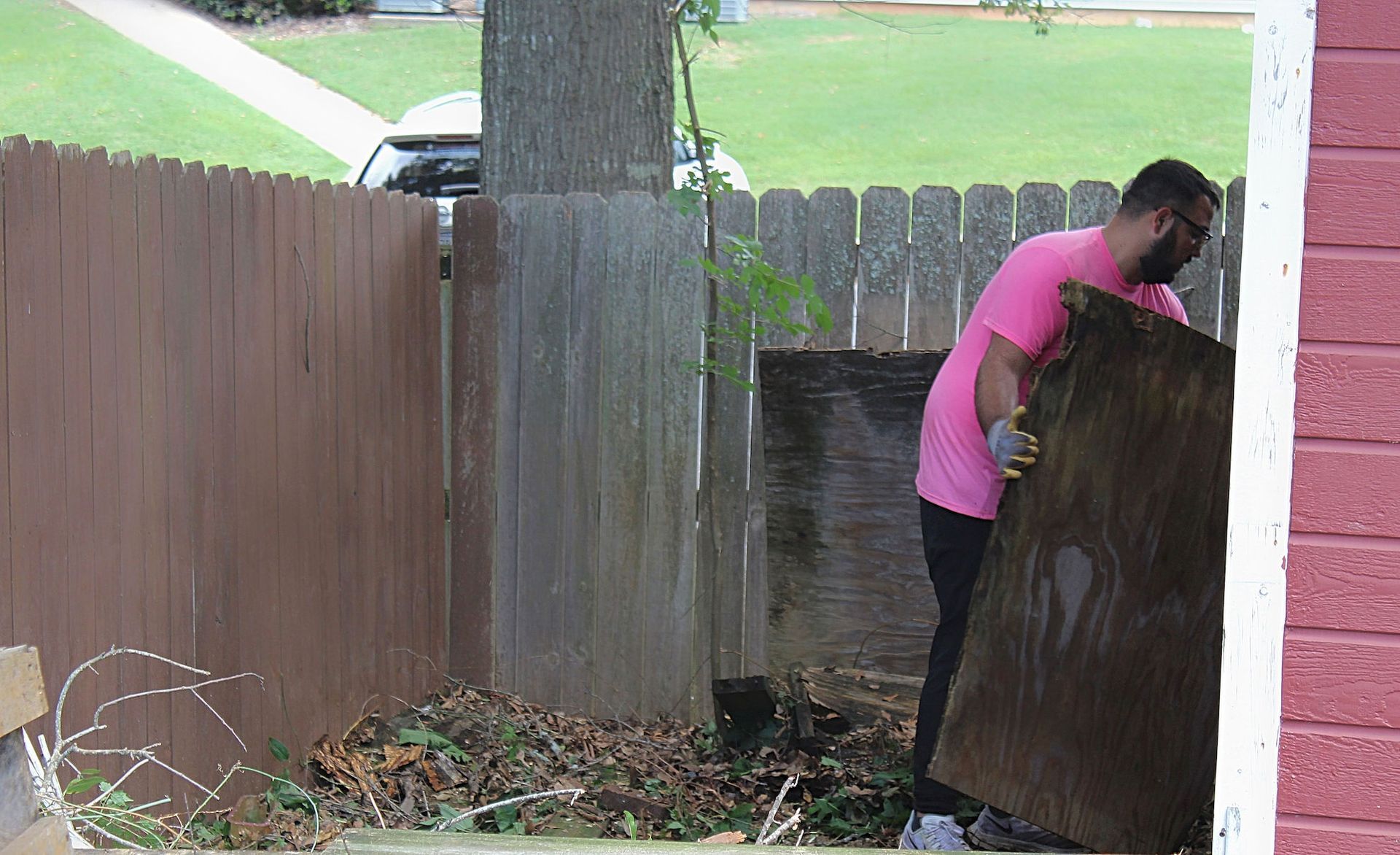 Two people moving a large brown fence panel beside a wooden gate in a yard