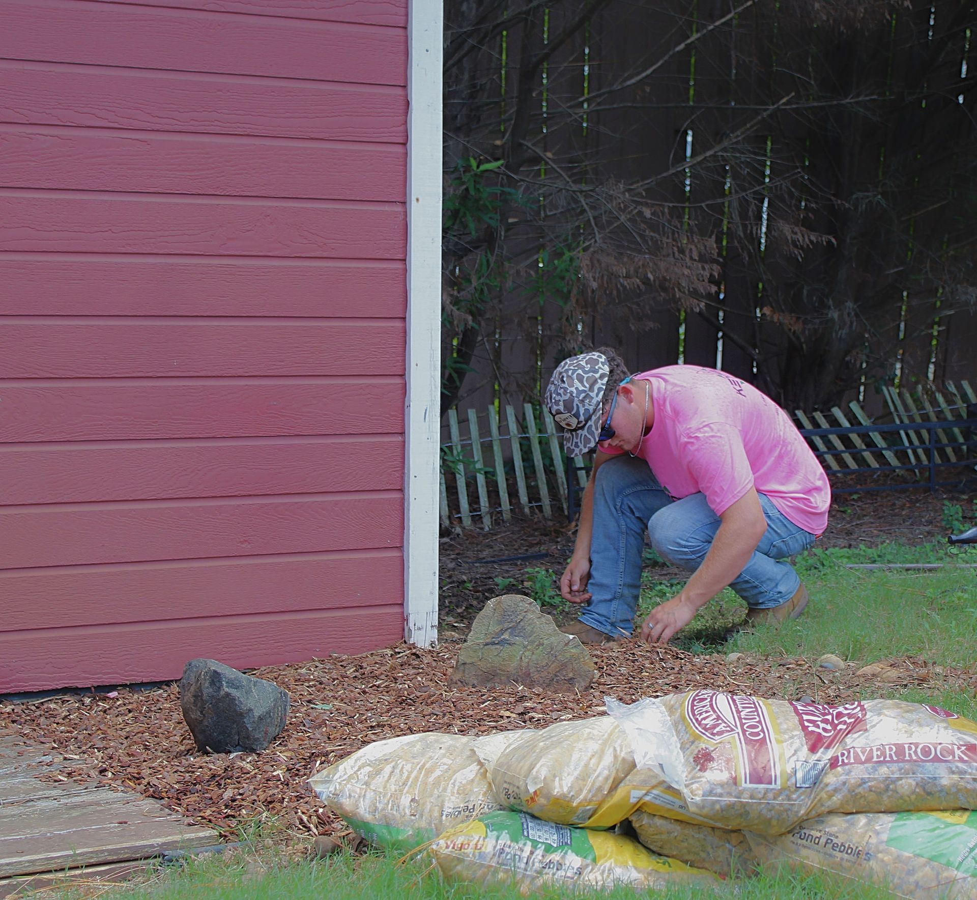 Person in a pink shirt crouches by a red building, digging in the garden beside landscaping supplies.