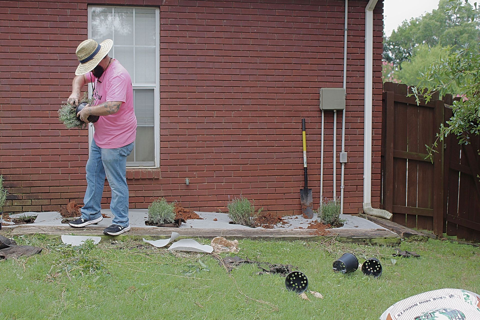 Person in pink shirt and straw hat gardening beside a brick house, holding a small dog.