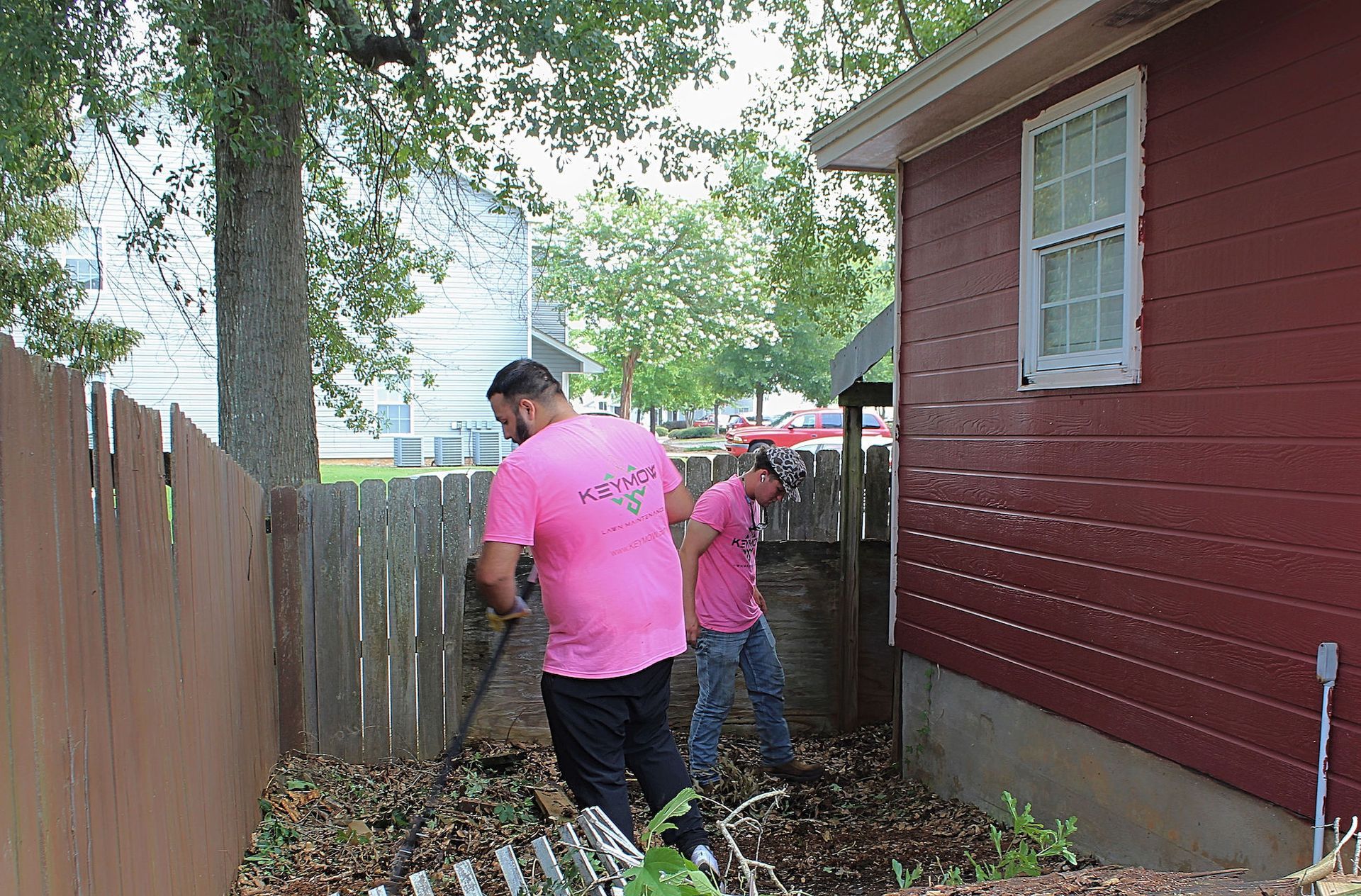 Two workers in pink shirts clear debris along a narrow side yard beside a red house and wooden fence