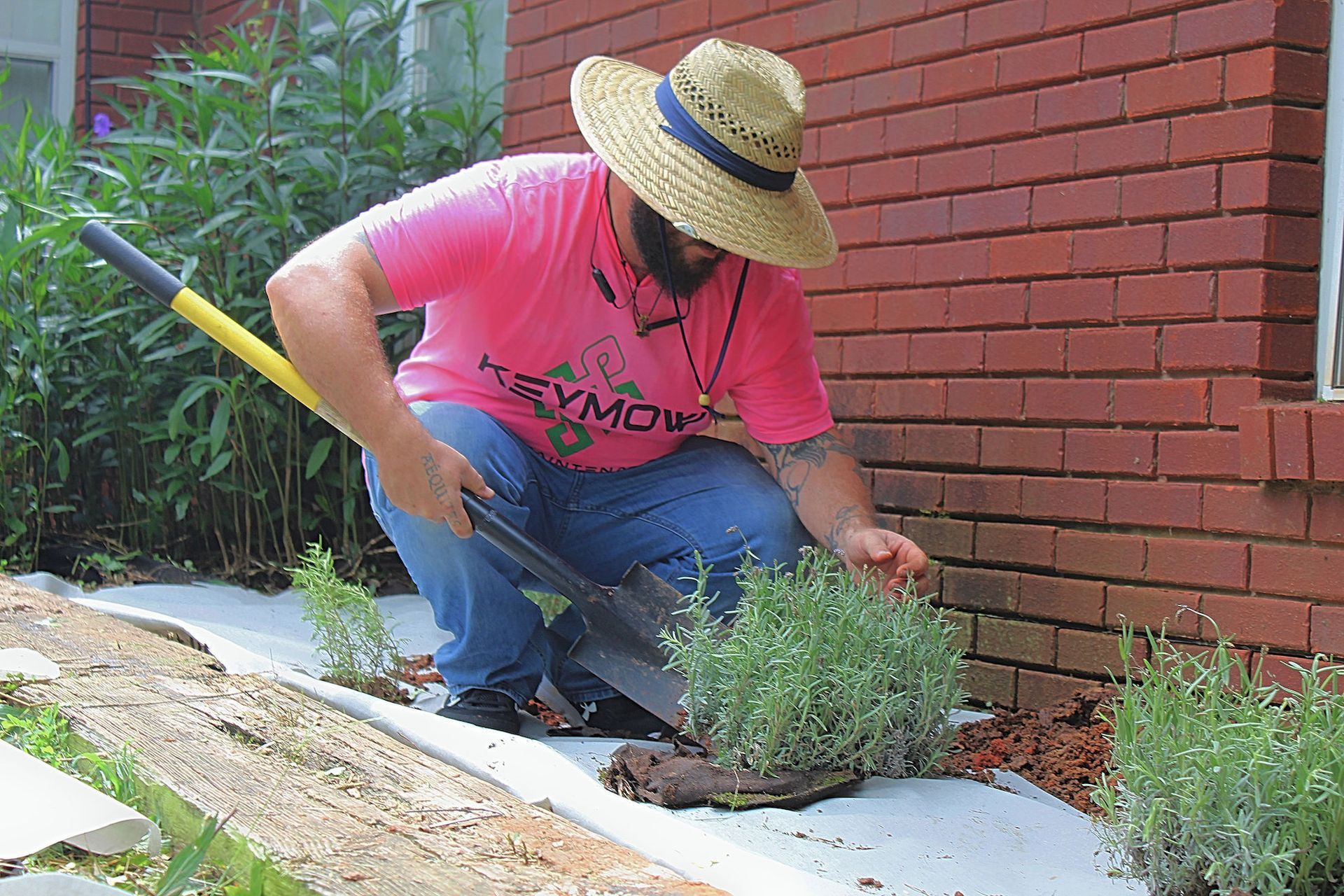Person in a straw hat kneeling by a brick wall, gardening plants in a flower bed.