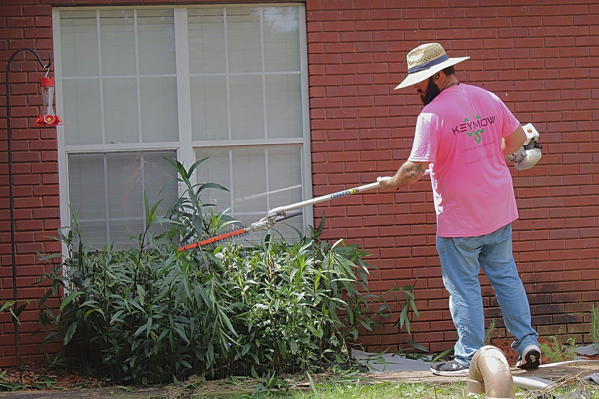 Person using a weed trimmer beside a brick house, cutting tall bushes near a window.
