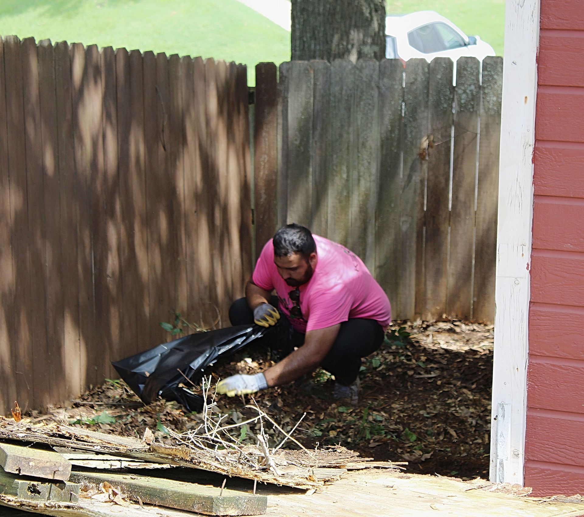 Person in a pink shirt kneels by a fence, gathering yard debris into a black bag.